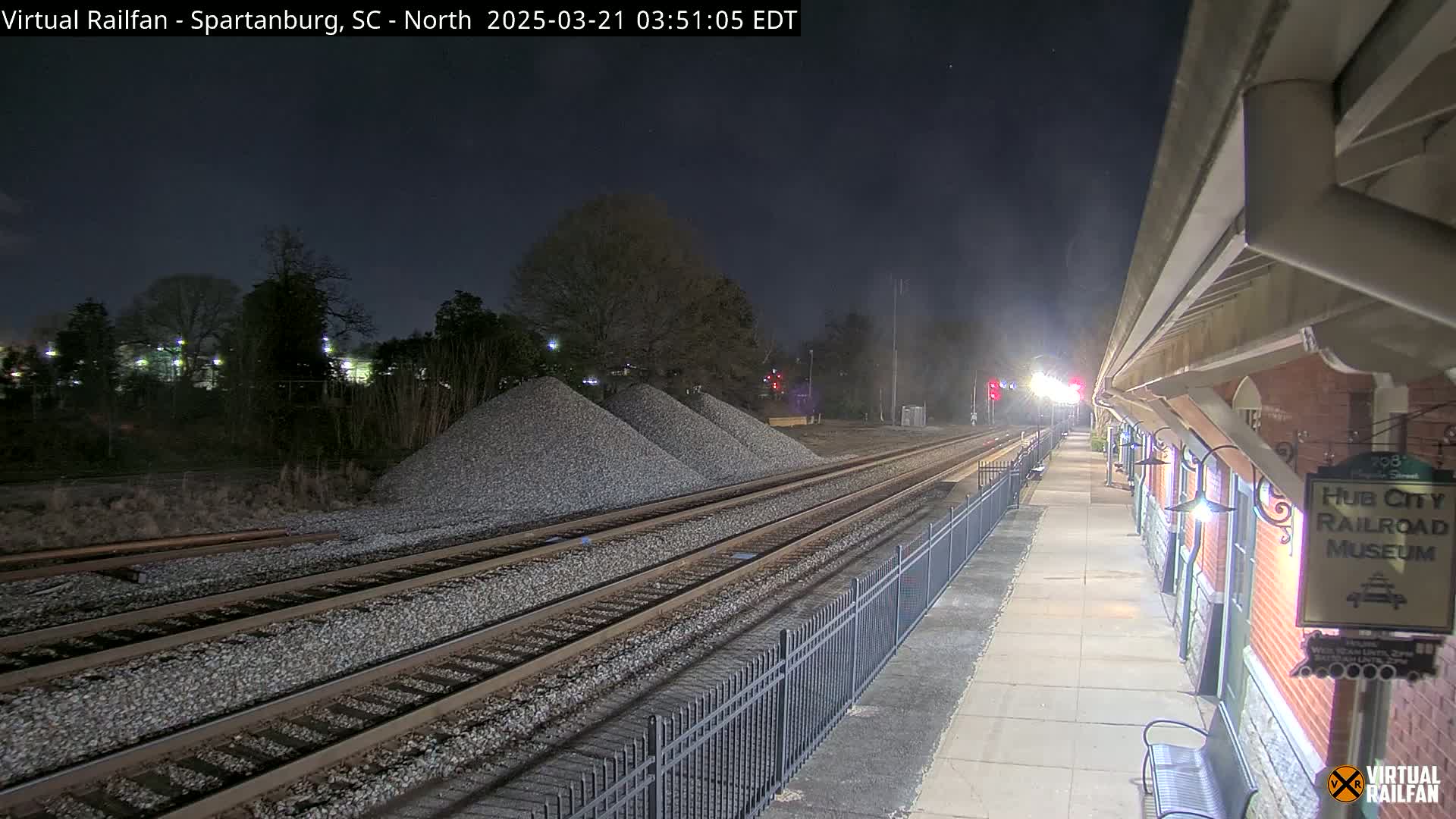 At night, a train station platform extends alongside railroad tracks that pass piles of gravel, under a clear, dark sky.