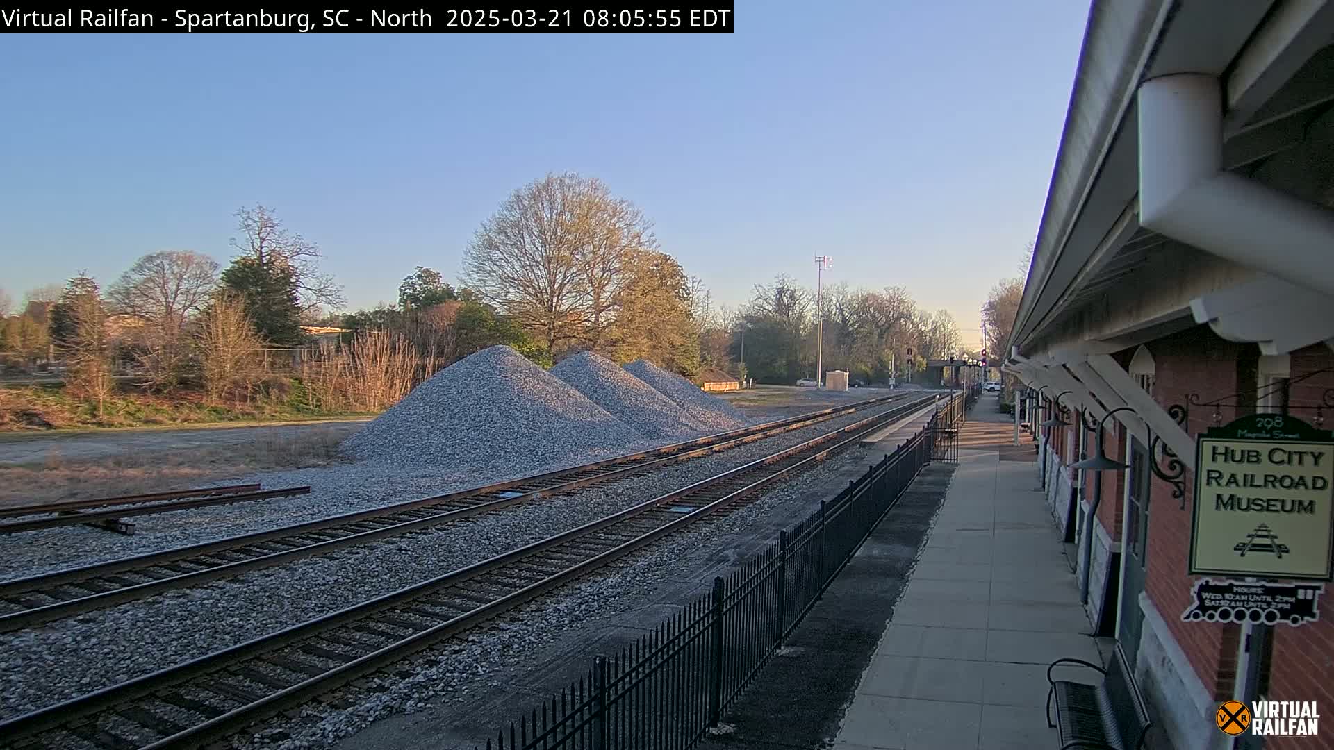 A train platform beside several piles of gravel and train tracks extends into the distance under a clear, bright sky.