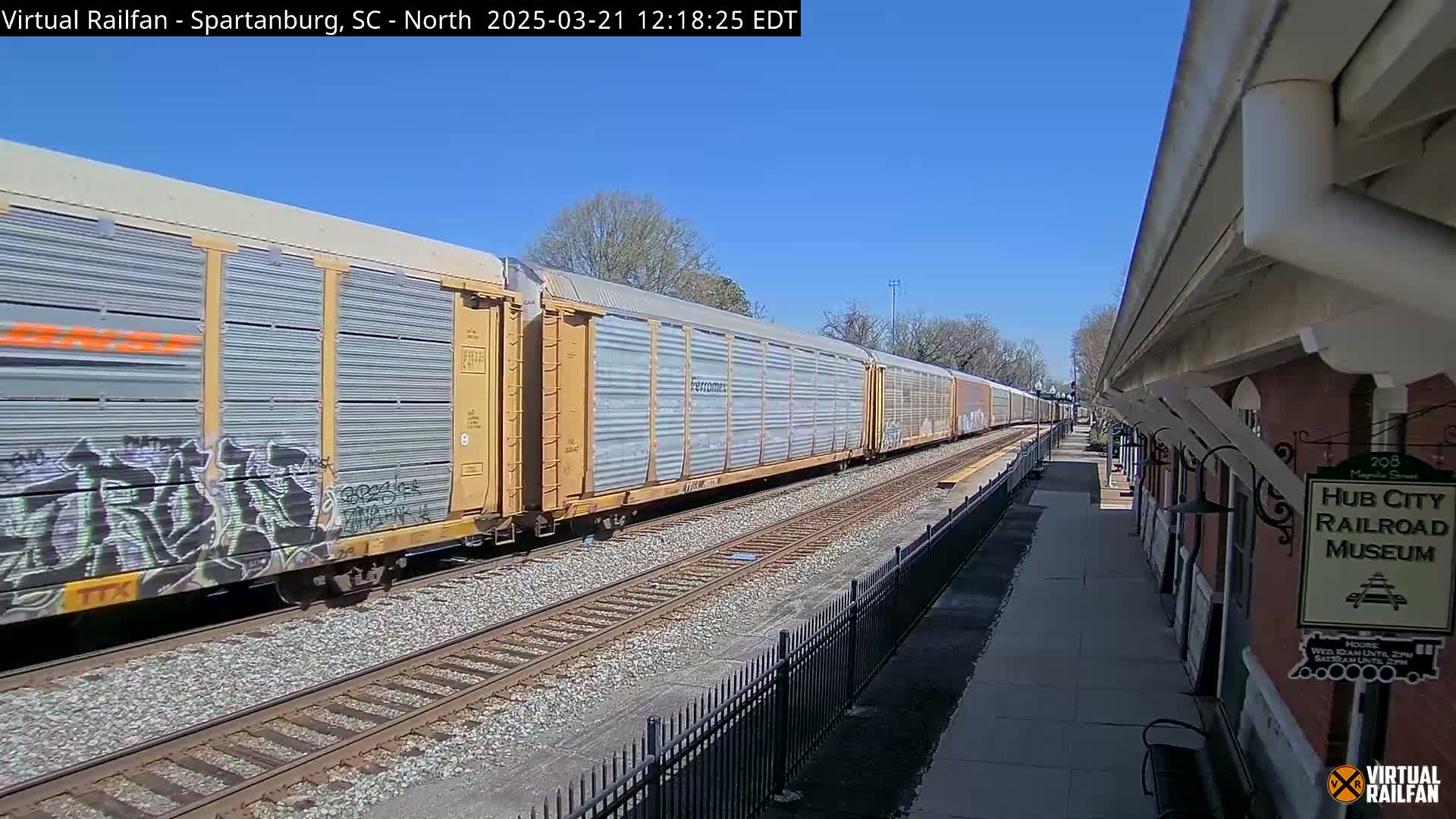 A long freight train with graffiti on some cars passes by a railroad museum on a sunny day.