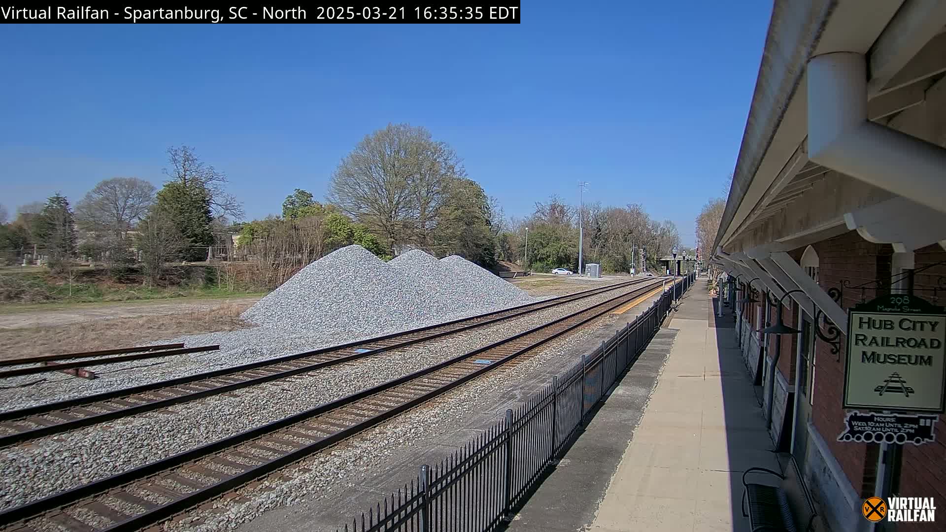 Under a clear blue sky, railroad tracks extend toward a brick building beside piles of gravel, with a black metal fence separating the tracks from the platform.