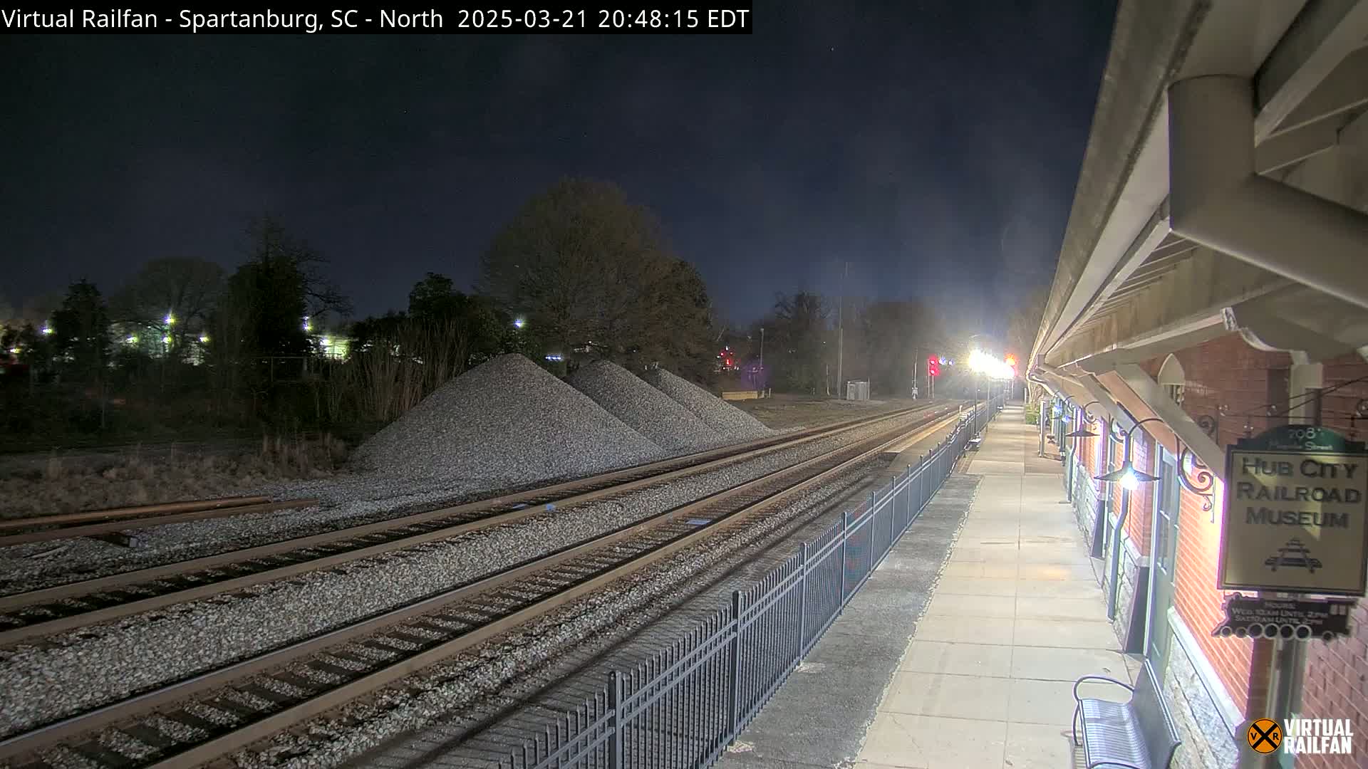 Under a dark night sky, train tracks extend from a nighttime train station platform with several piles of gravel nearby.