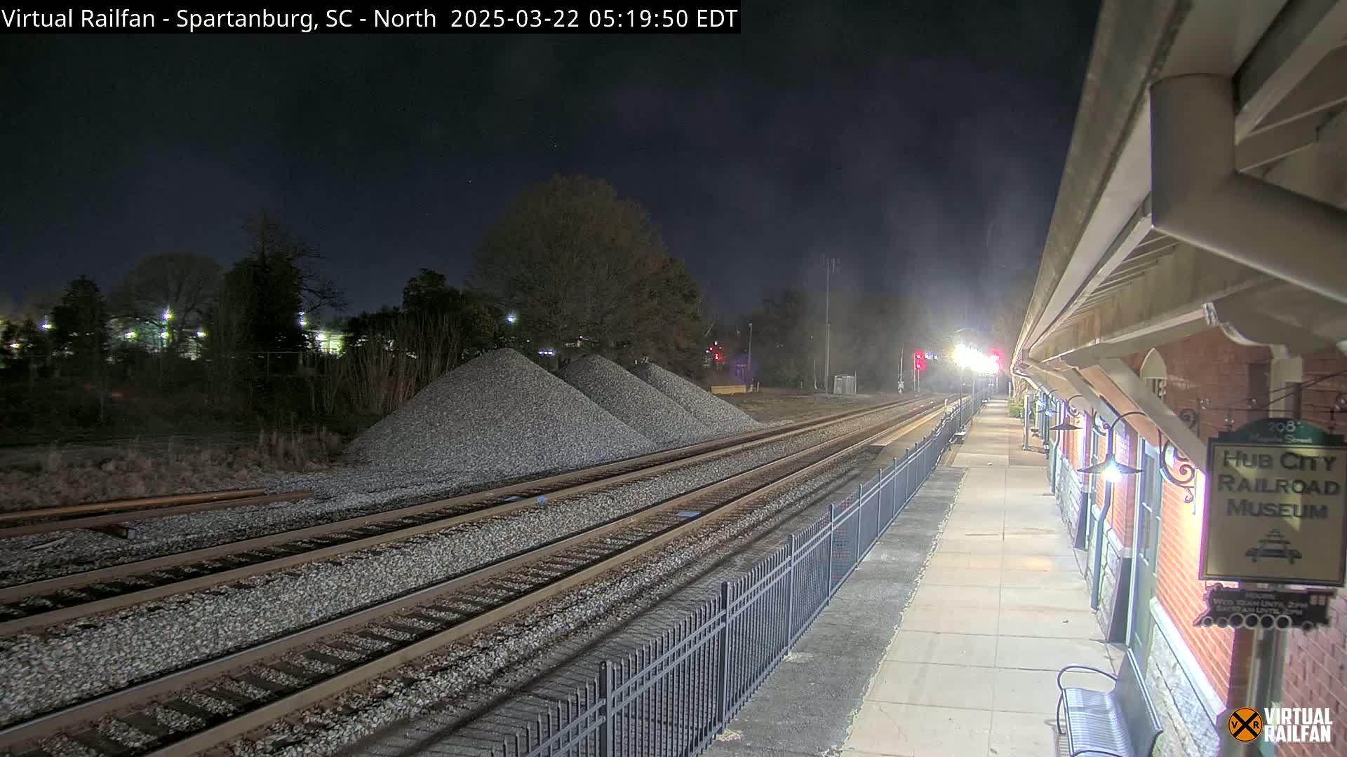 At night, railroad tracks extend from a train platform next to a brick building, with piles of gravel nearby under a clear, dark sky.