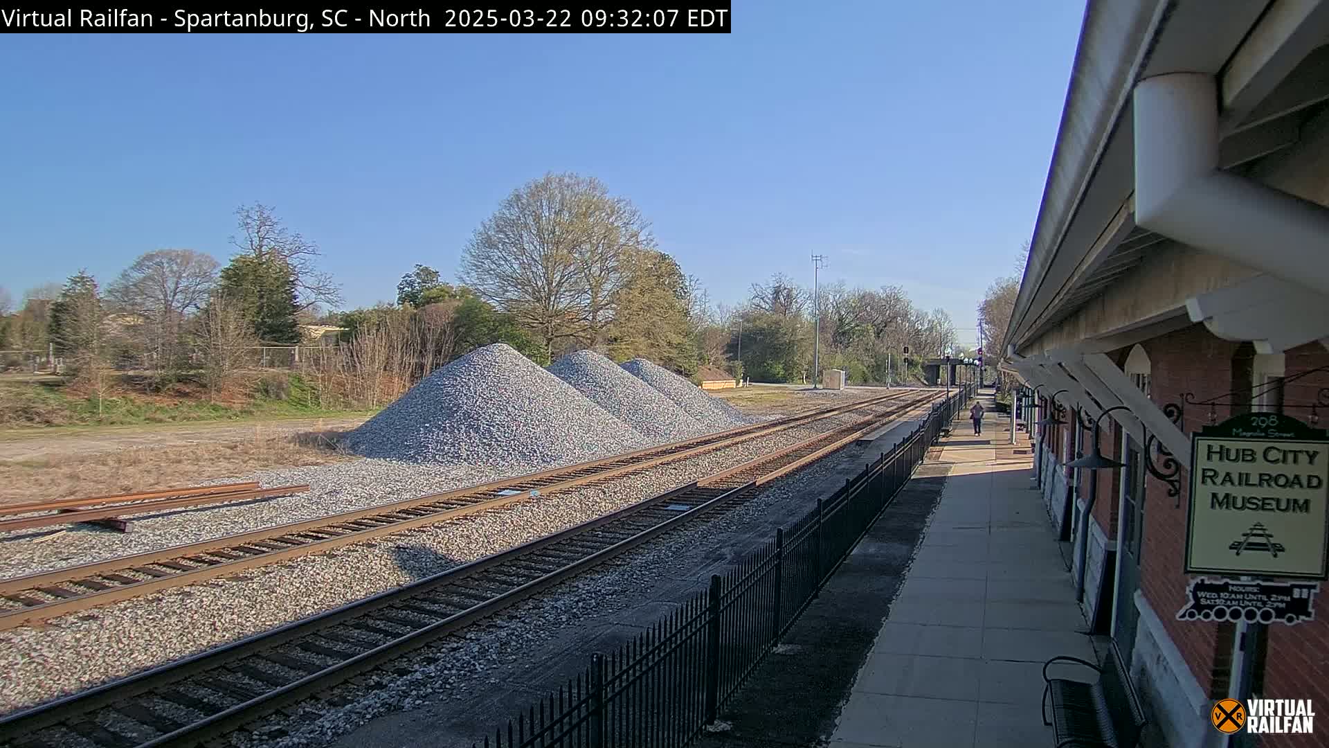 Under a clear blue sky, railroad tracks run alongside a brick building and several piles of gravel, with a person walking in the distance.
