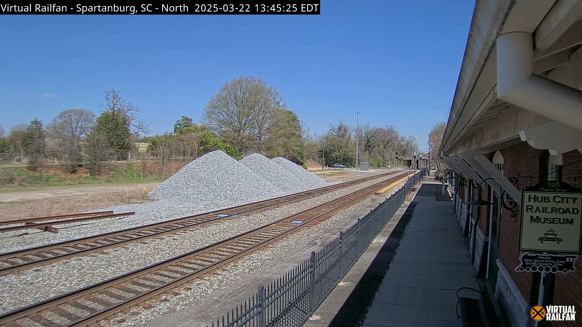 A sunny day reveals railroad tracks beside a depot, with piles of gravel nearby.