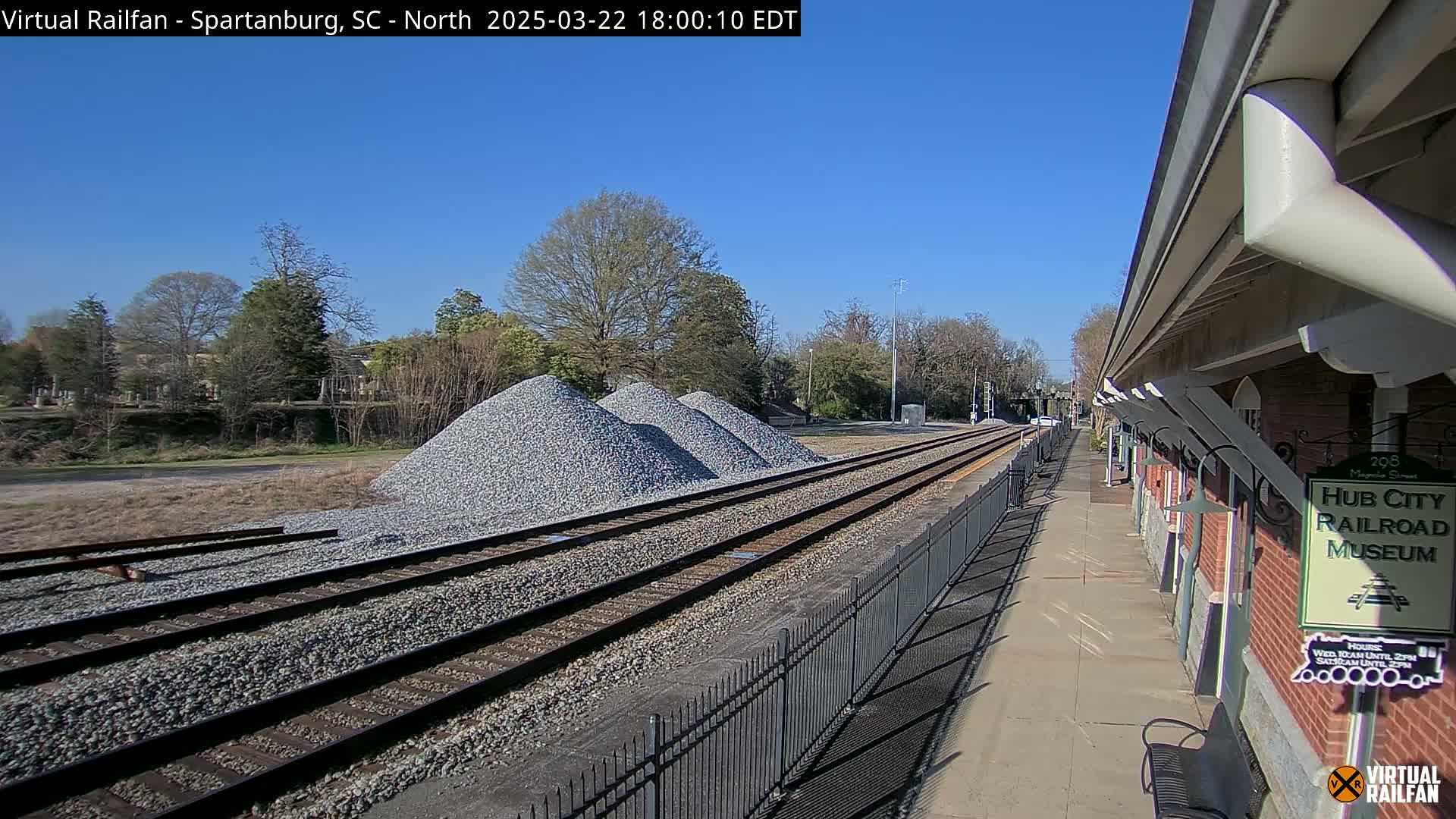 Under a clear blue sky, railroad tracks extend from a train platform beside piles of gravel toward a tree-lined horizon.