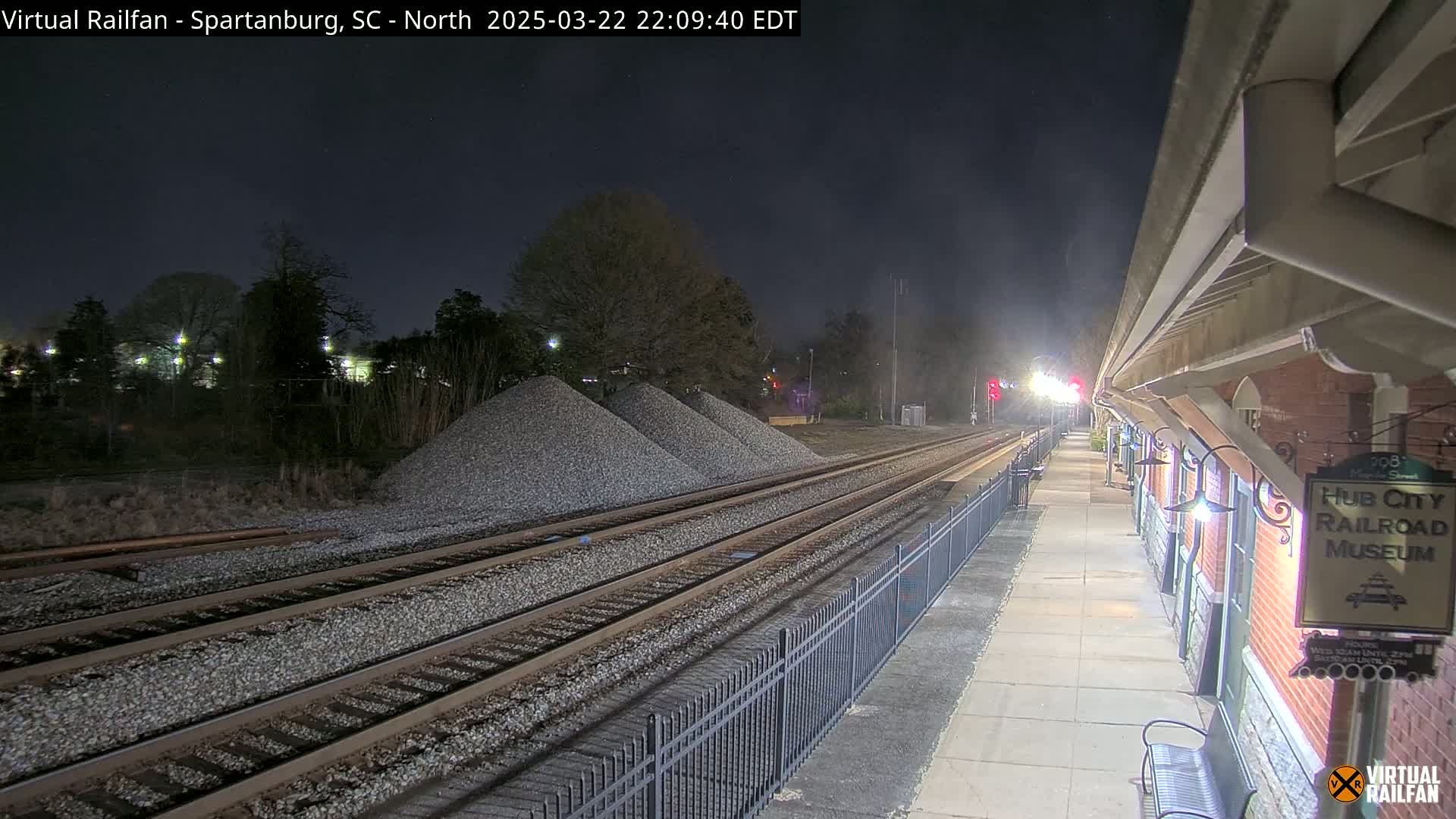 At night, a train platform next to railroad tracks with piles of gravel extends into the distance under a dark sky.