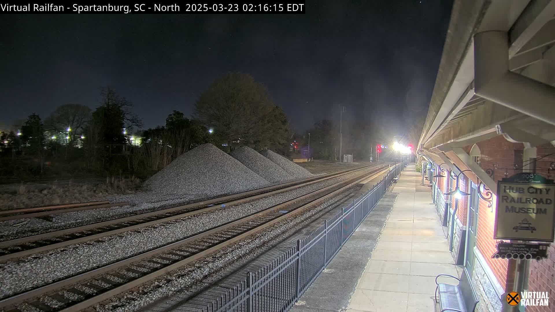 A nighttime view of train tracks, with piles of gravel nearby, leading to a brick railroad museum platform under a clear, dark sky.