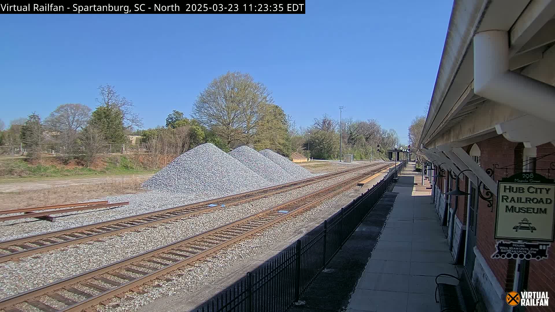 Under a clear blue sky, railroad tracks extend alongside a brick building and several piles of gravel.
