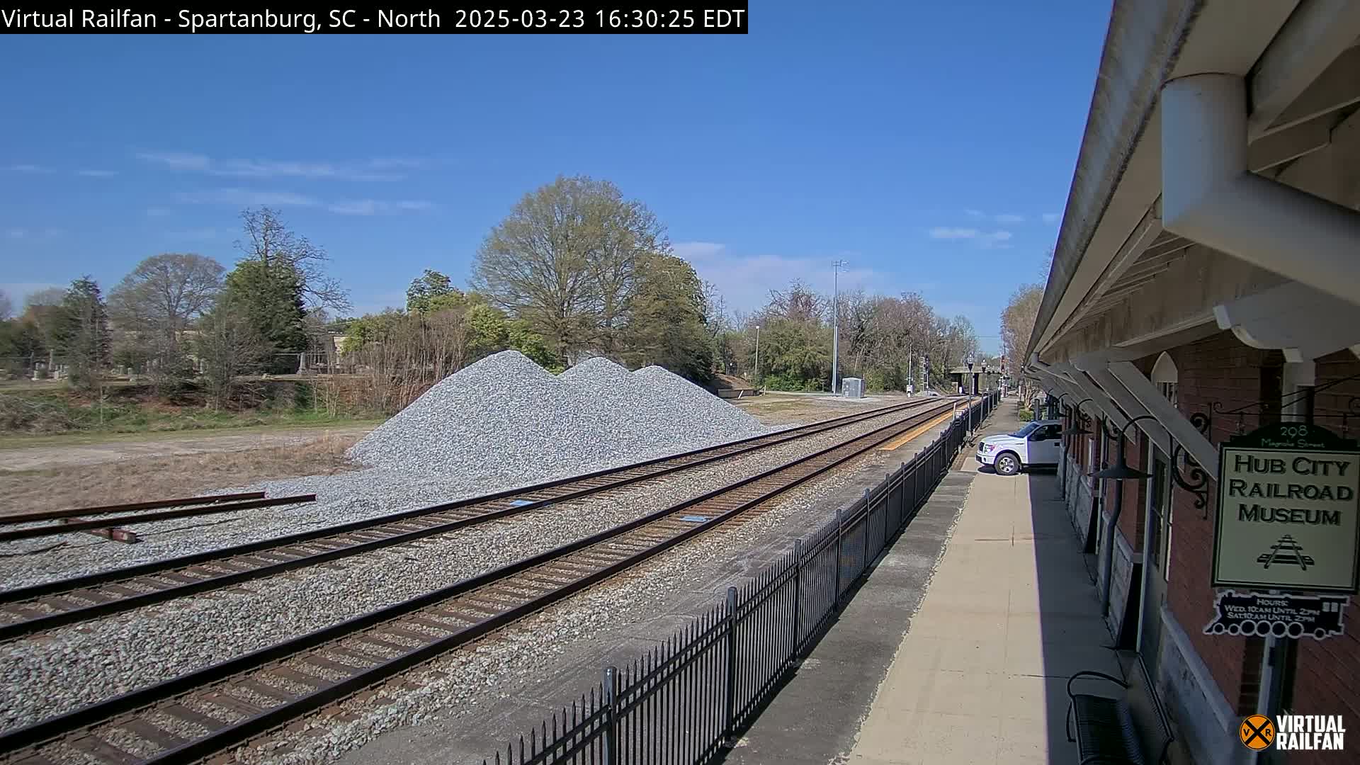 A sunny day reveals train tracks alongside a brick building, a gravel pile, and a parked truck.