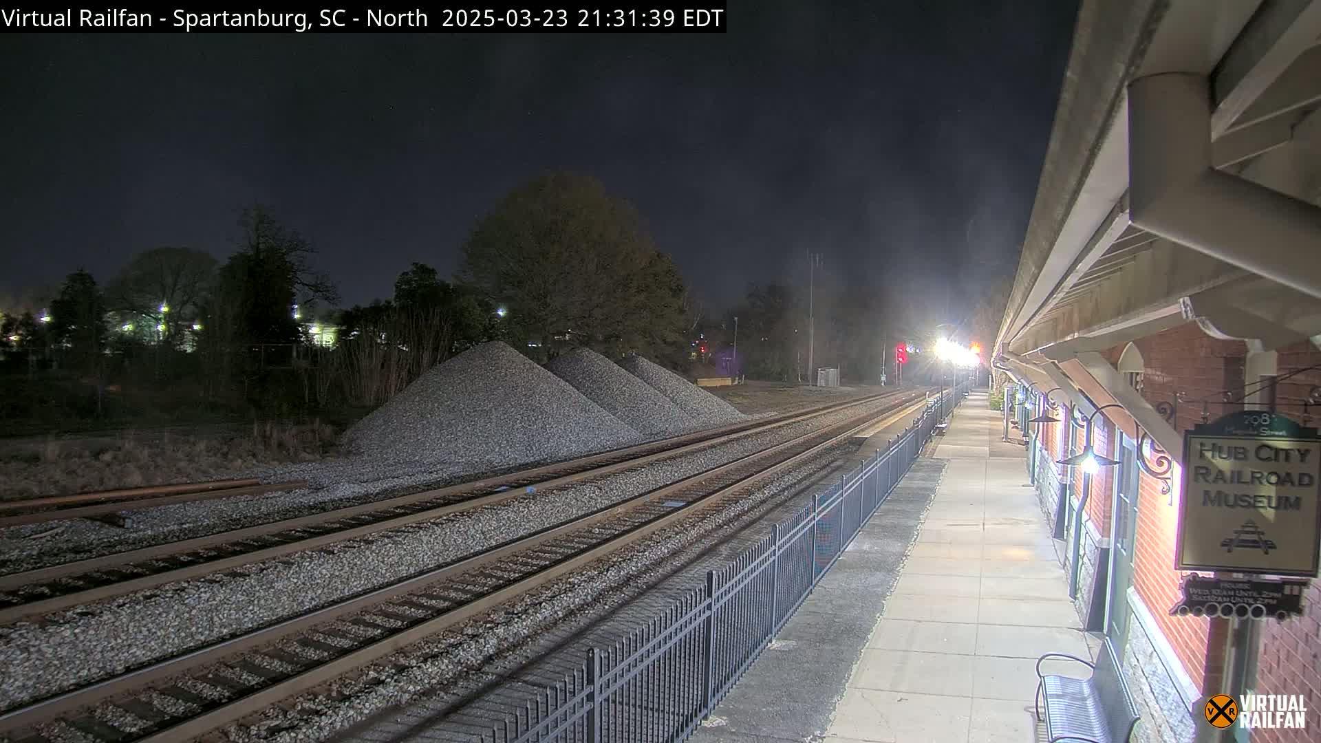 A nighttime view of train tracks leading to a brick railroad museum building, with piles of gravel nearby under a dark sky.