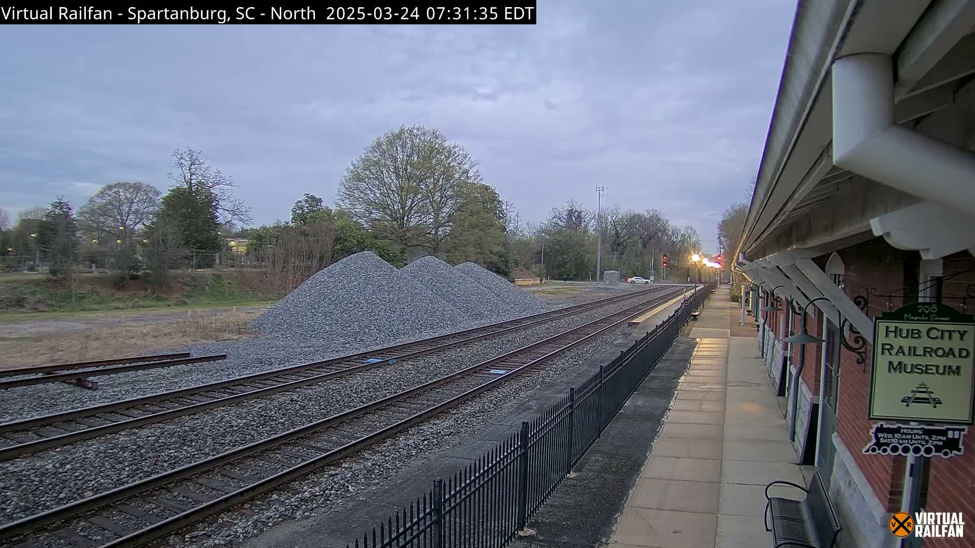 A train platform with a brick building and multiple train tracks extends into the distance under an overcast sky, next to piles of gravel.
