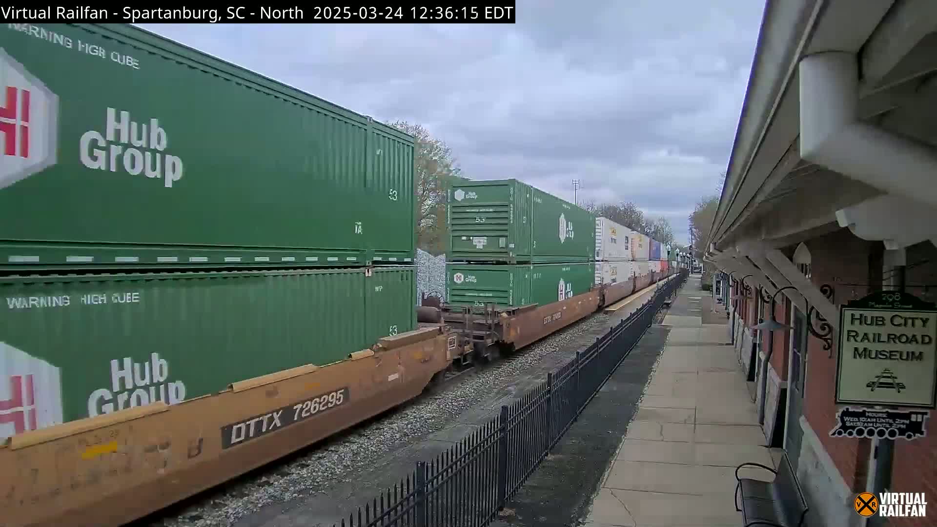 A freight train carrying green shipping containers passes by a railroad museum on an overcast day.