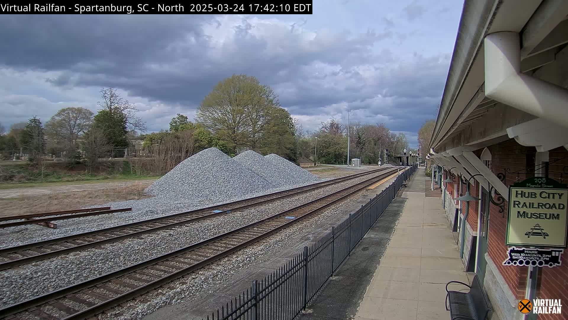 A train platform next to railroad tracks with piles of gravel, under a partly cloudy sky.