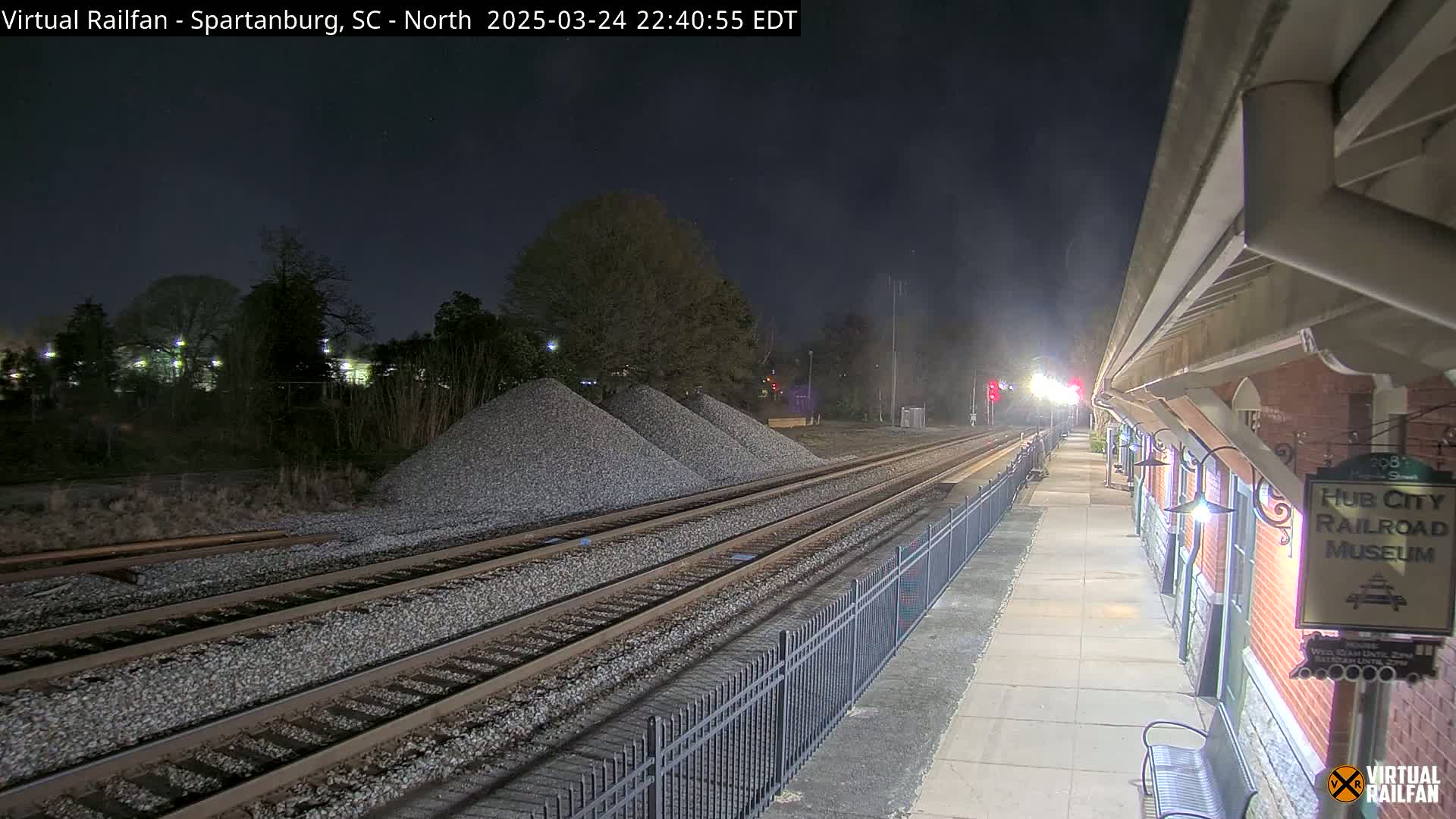 At night, railroad tracks extend from a train platform beside a brick building, past piles of gravel under a dark sky.