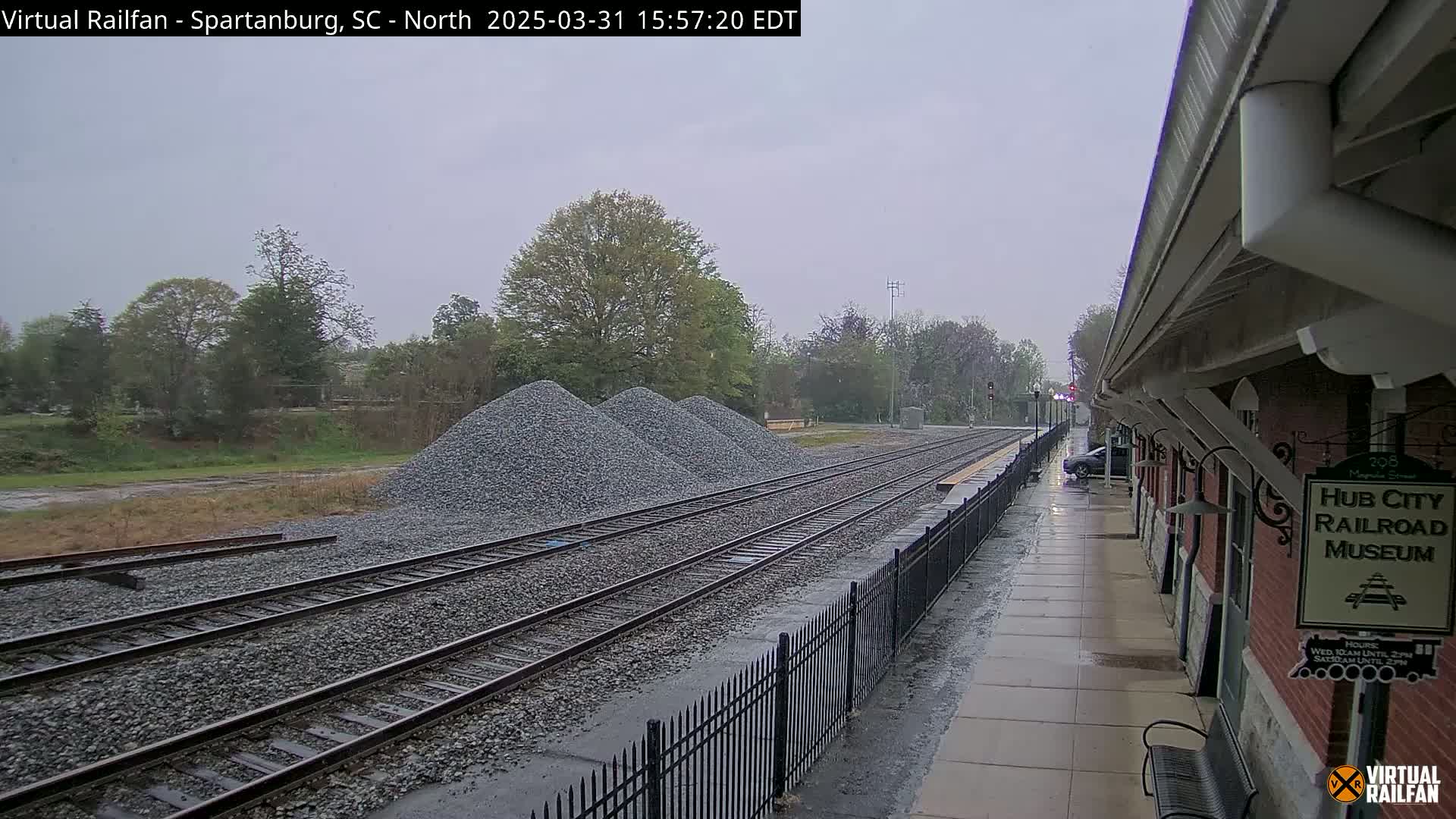 A rain-slicked railway platform next to a brick building overlooks several piles of gravel beside multiple train tracks.