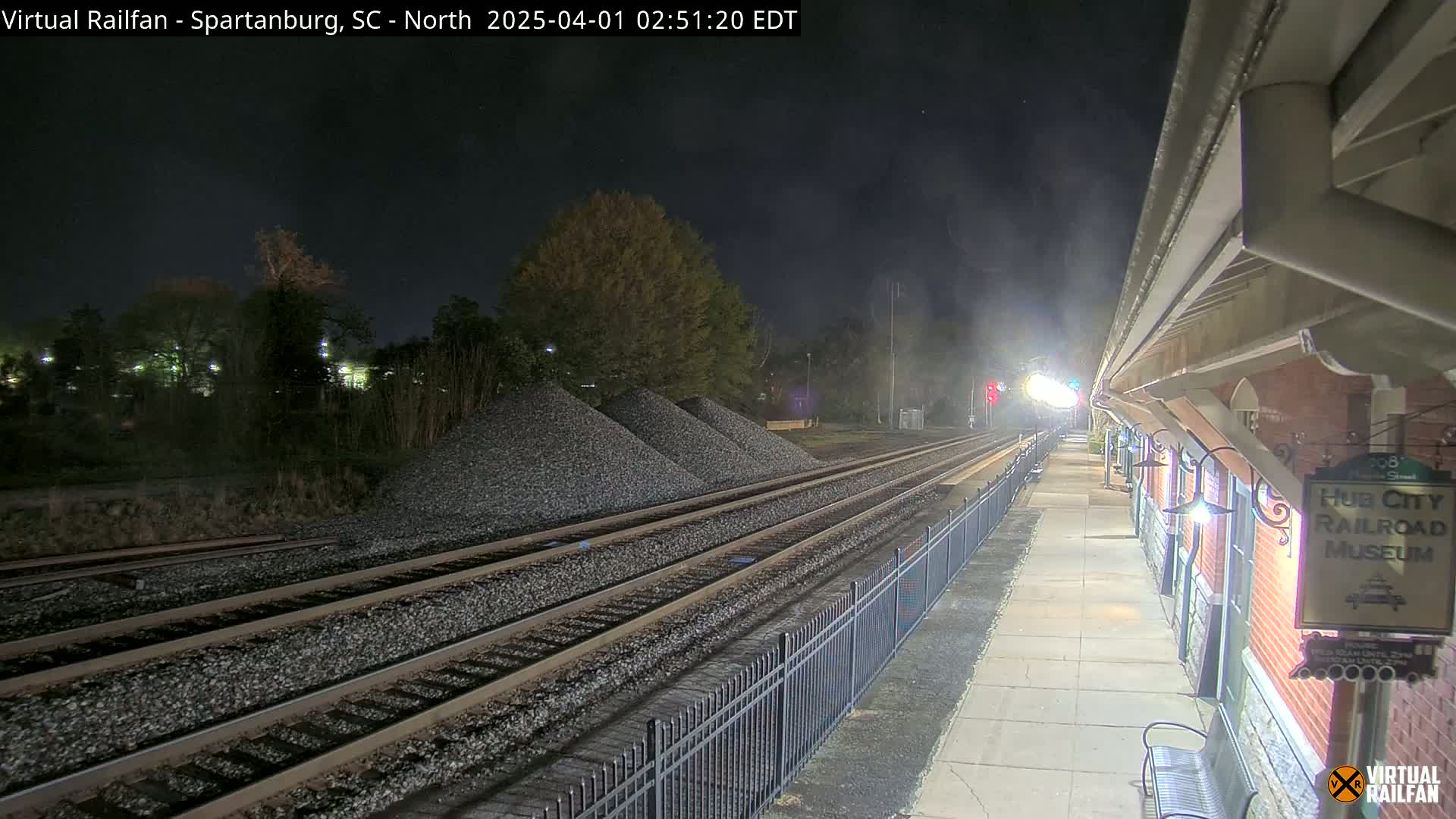 At night, several train tracks extend from a brick railroad museum platform towards piles of gravel under a dark sky.