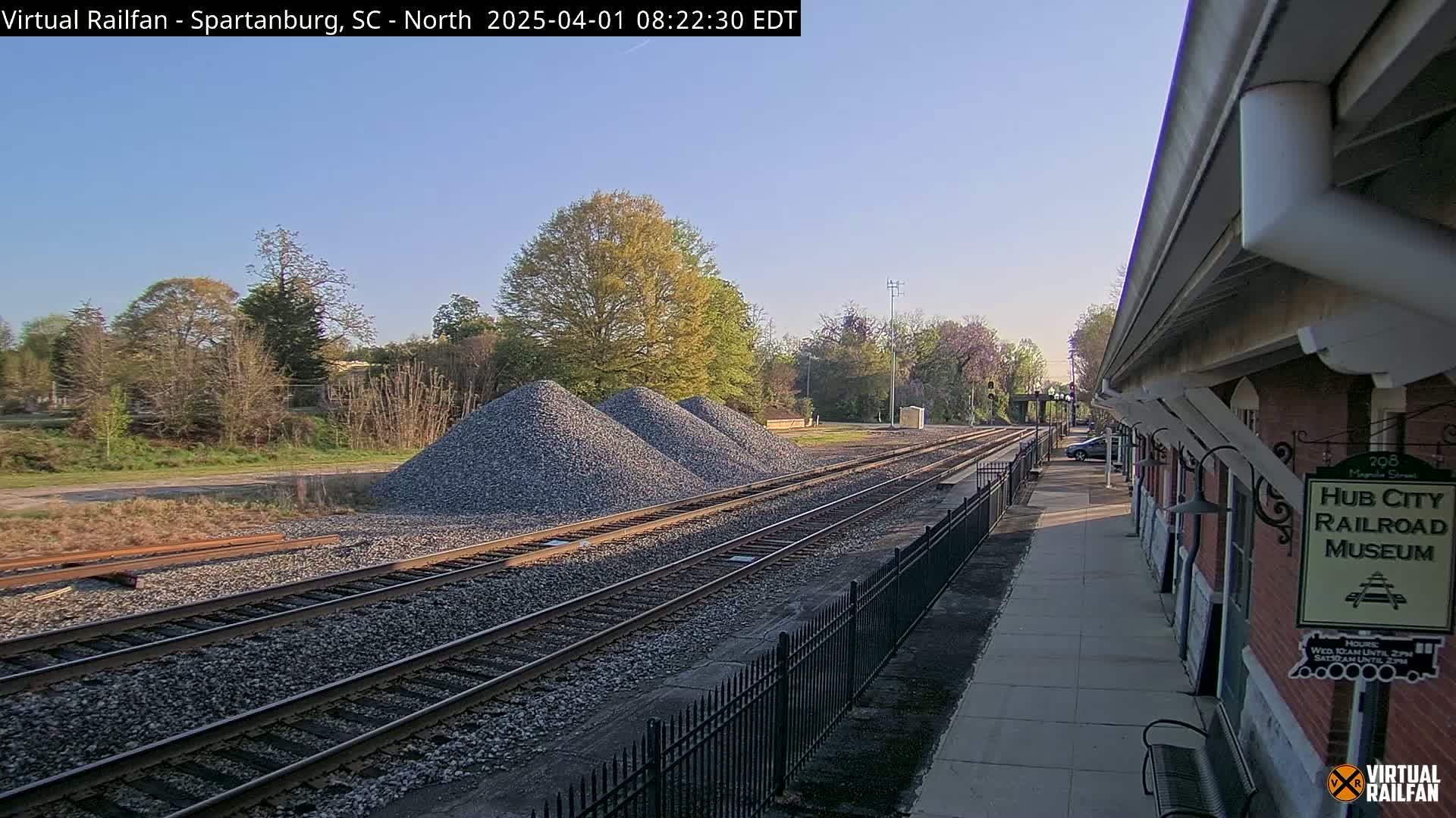 Under a clear blue sky, railroad tracks extend from a brick railroad museum building toward piles of gravel and a treeline.