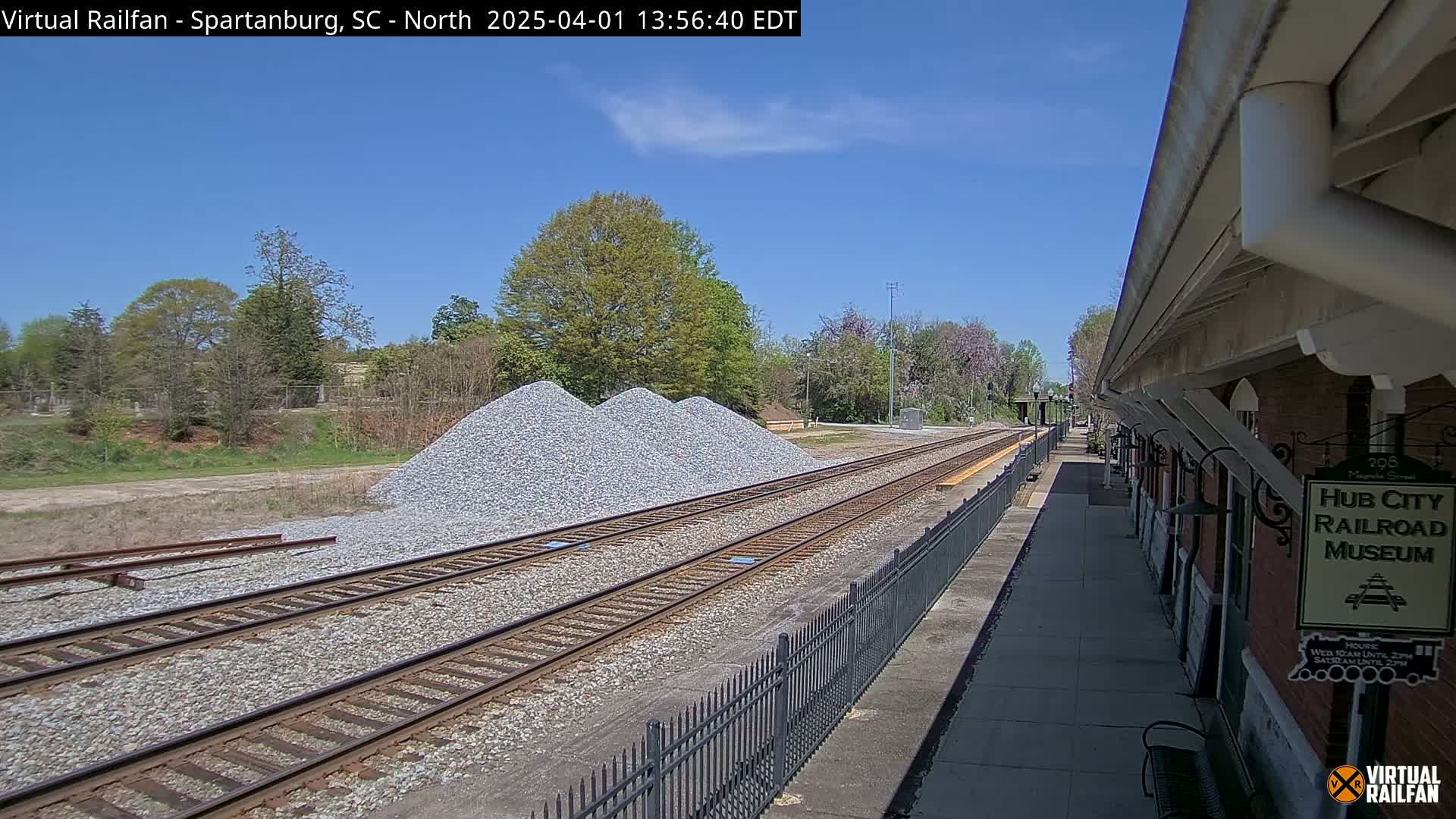 A sunny day reveals railroad tracks extending from a train platform beside a brick building next to piles of gravel under a clear blue sky.