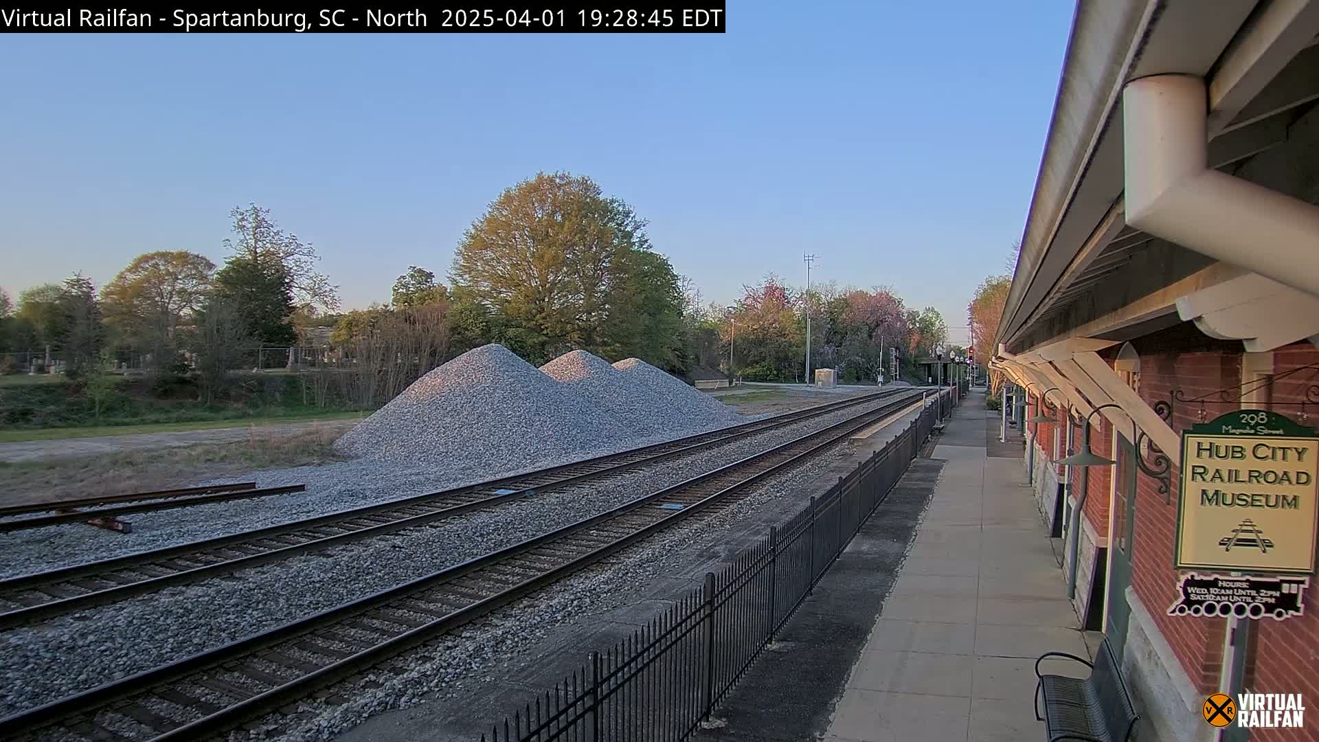 Under a clear twilight sky, railroad tracks extend from a brick railroad museum toward piles of gravel beside a chain-link fence.