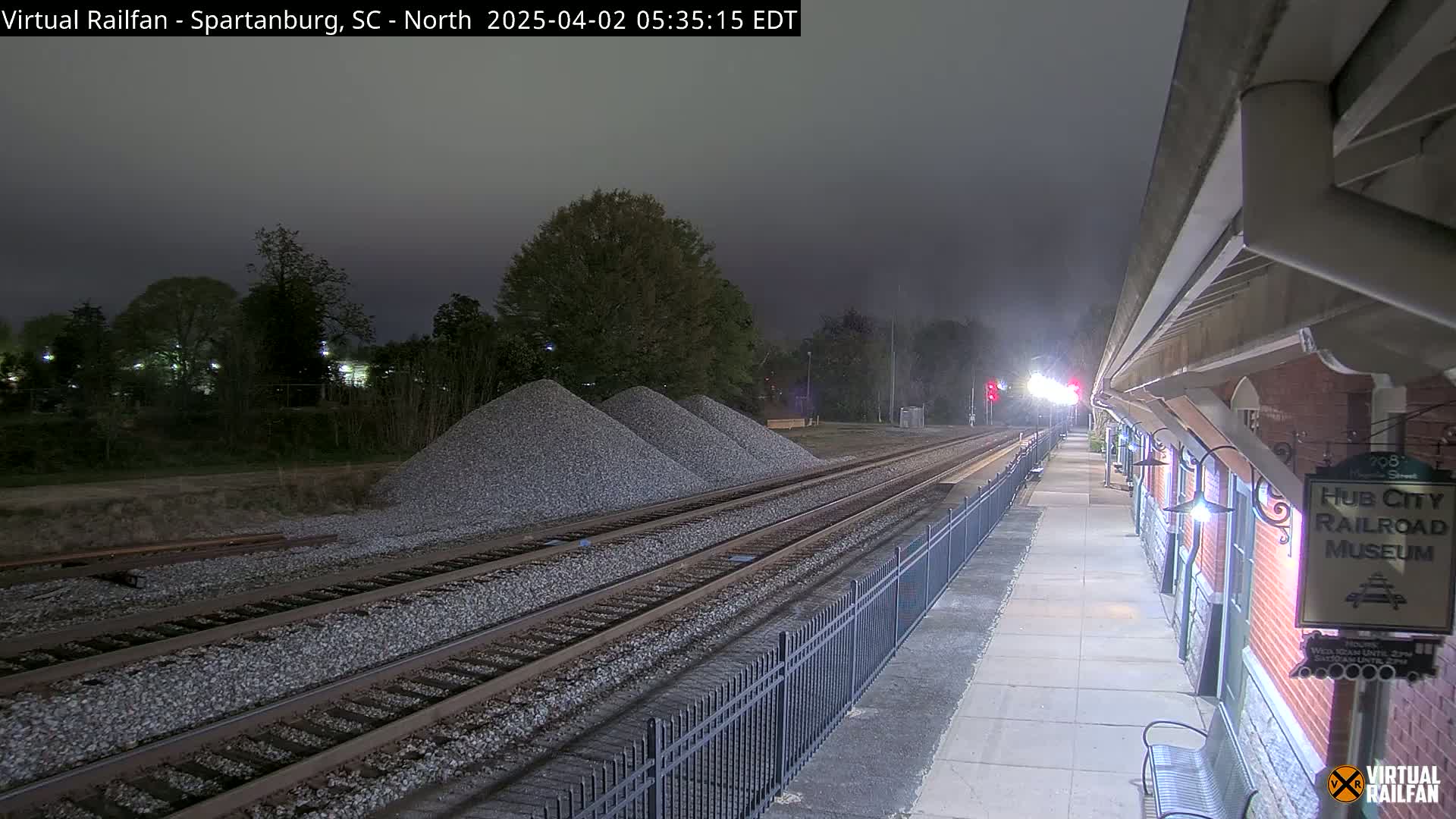 Under a dark, overcast night sky, multiple train tracks extend toward a brick building next to piles of gravel.