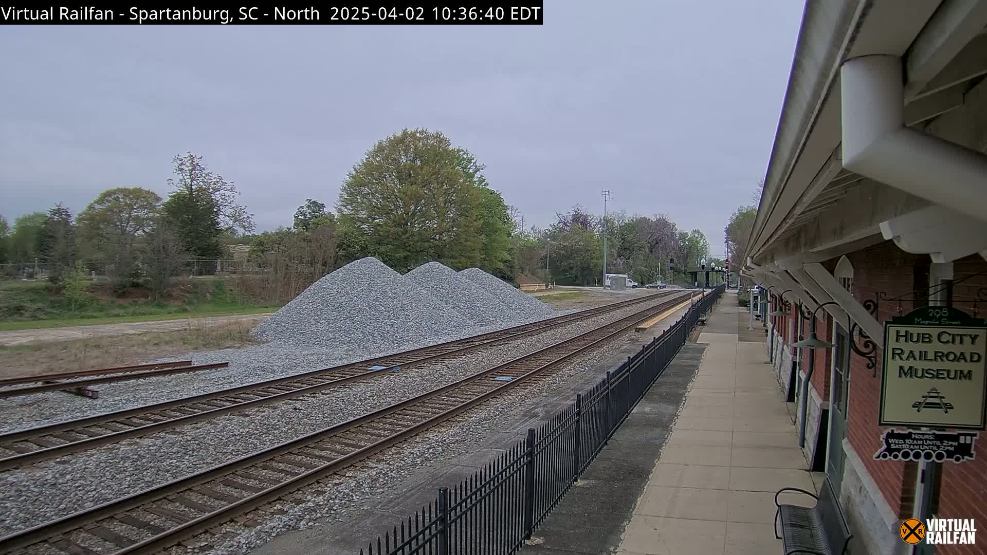 Overcast skies overlook railroad tracks with three piles of gravel beside them, a fenced platform, and a brick building.