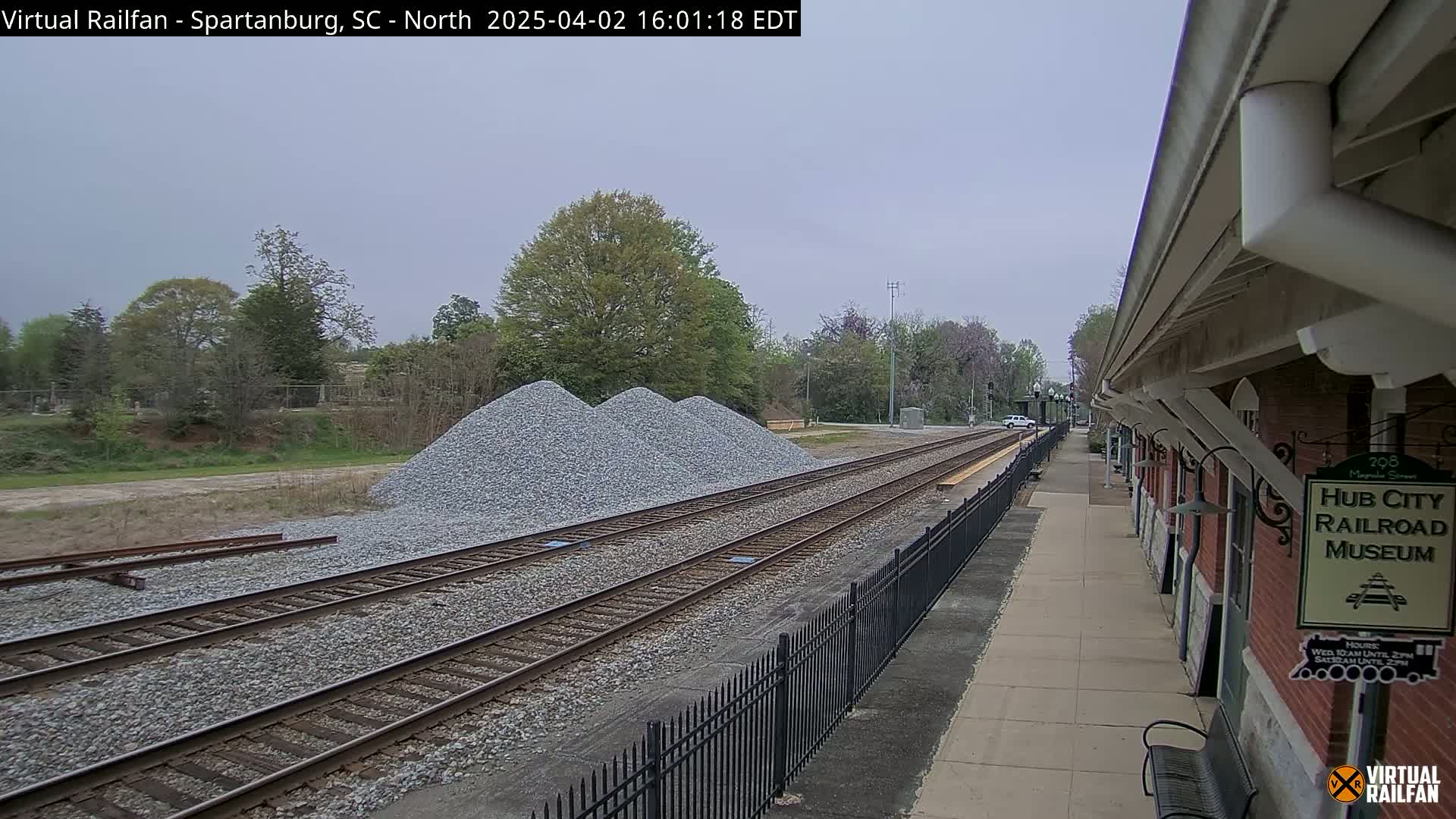 A train station platform next to railroad tracks with piles of gravel, under an overcast sky.