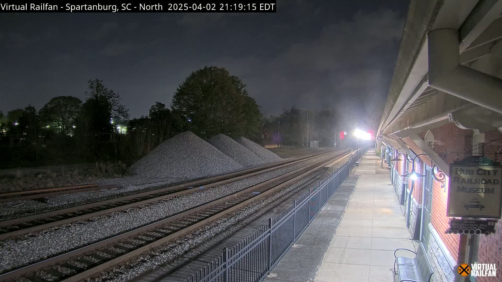 At night, train tracks extend from a nighttime railway station toward piles of gravel under a cloudy sky.