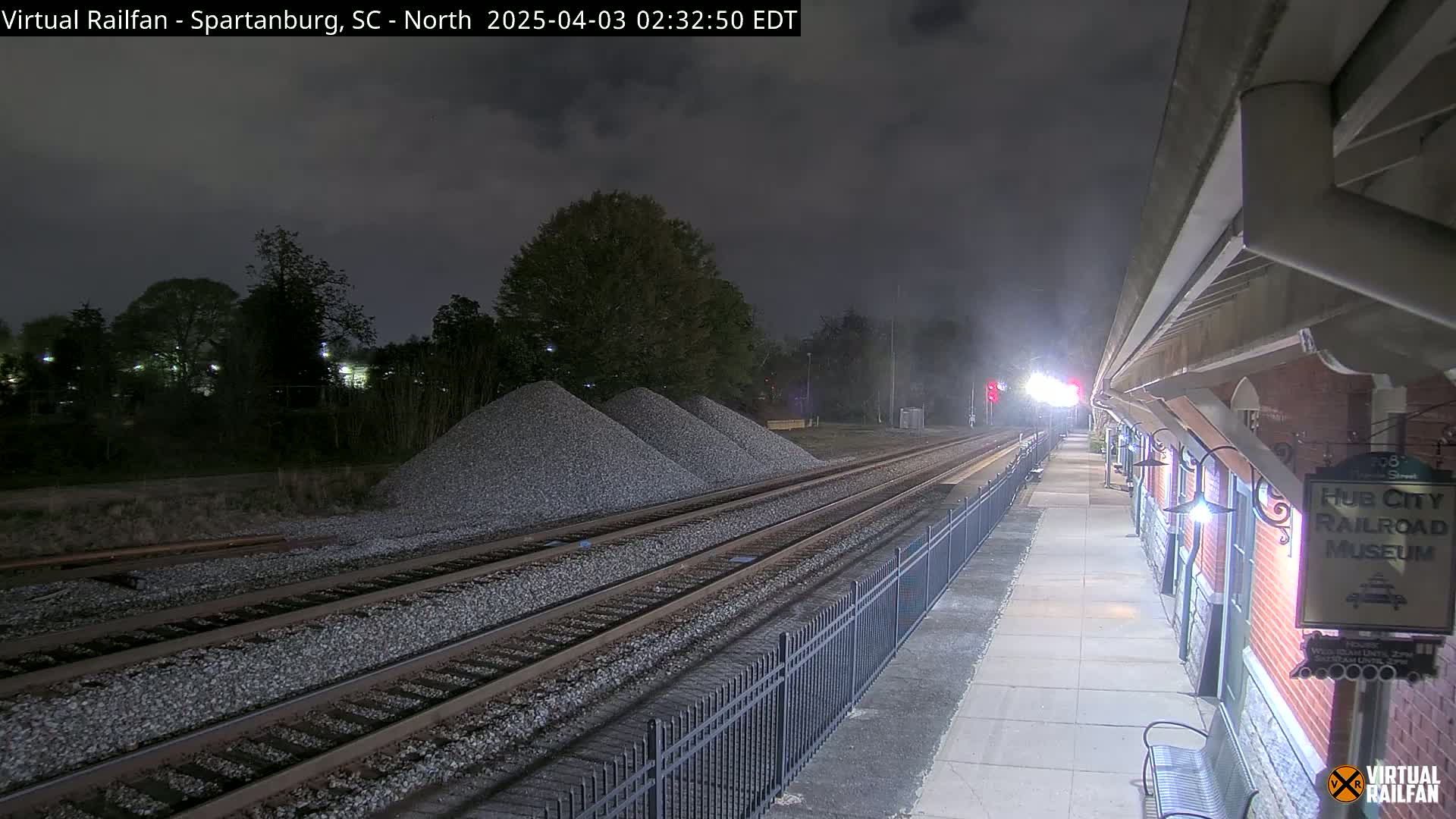 At night, under a cloudy sky, railroad tracks extend from a platform next to a brick building toward piles of gravel.