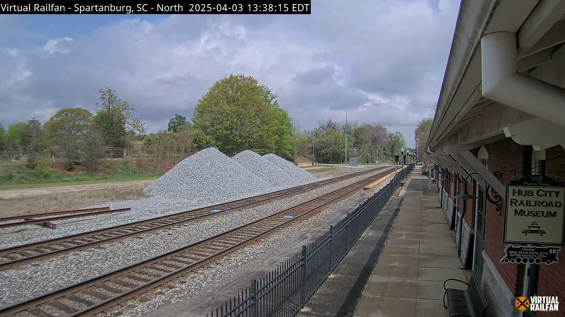 A train platform next to railroad tracks runs beside a brick building, with piles of gravel nearby under a partly cloudy sky.