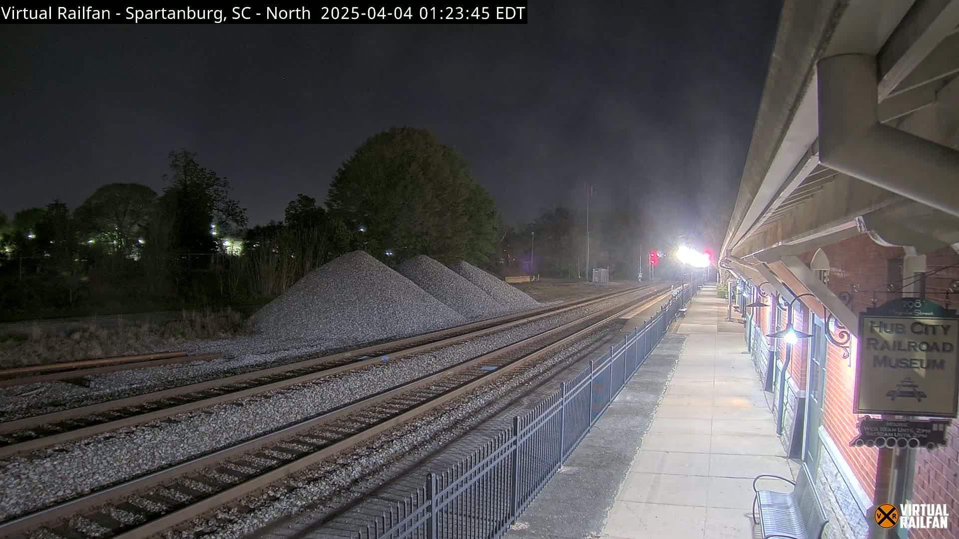 At night, a train station platform with a brick building is visible, next to railroad tracks with piles of gravel, under a dark, mostly clear sky.