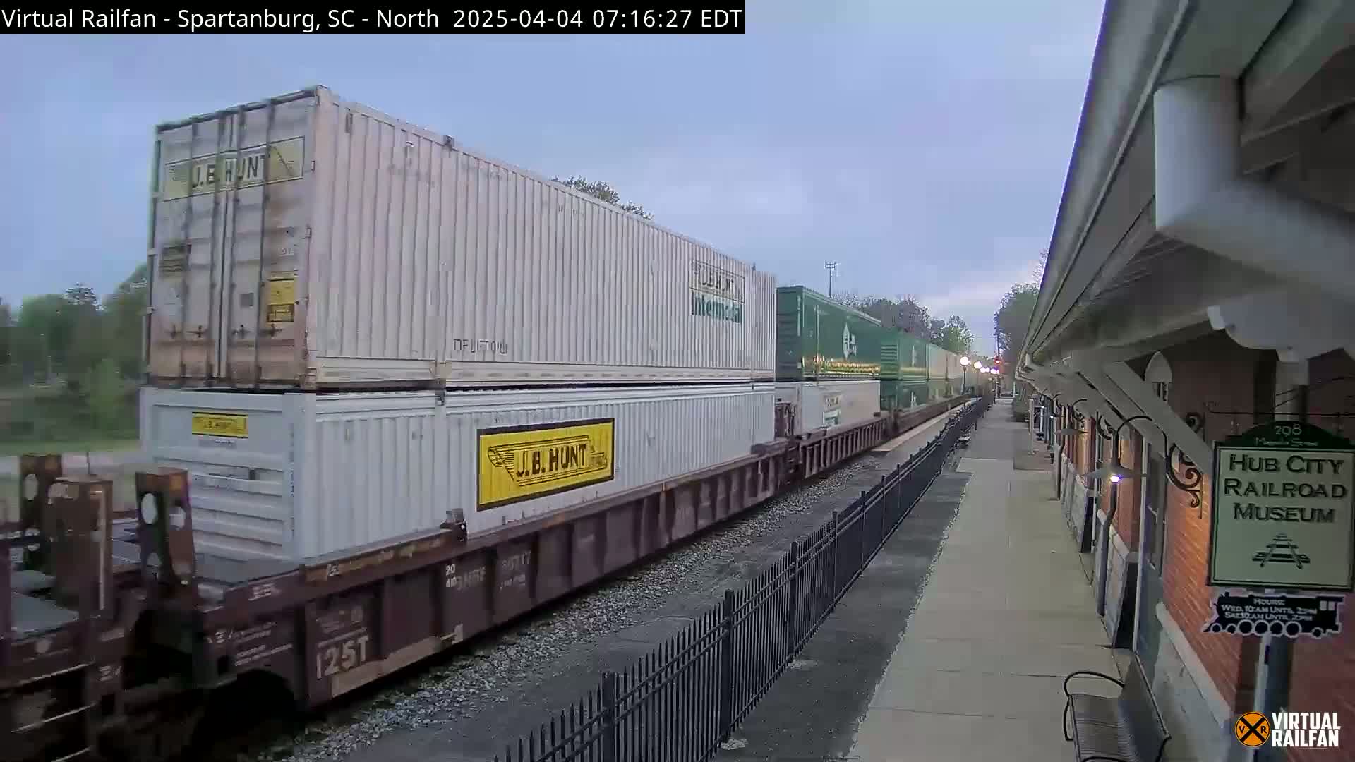 A freight train carrying stacked shipping containers passes a brick railroad museum building under an overcast sky.