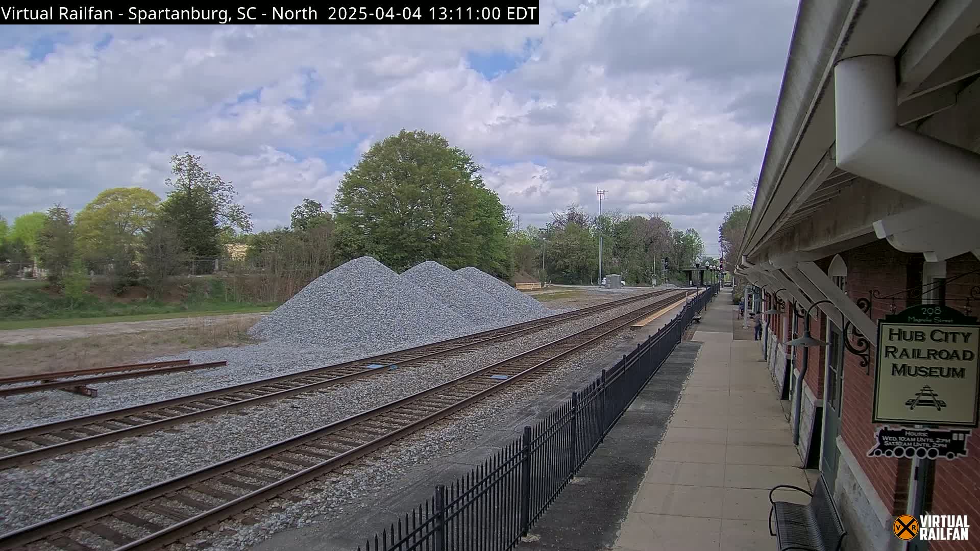 A partly cloudy day shows railroad tracks leading to a brick railroad museum, with piles of gravel nearby.