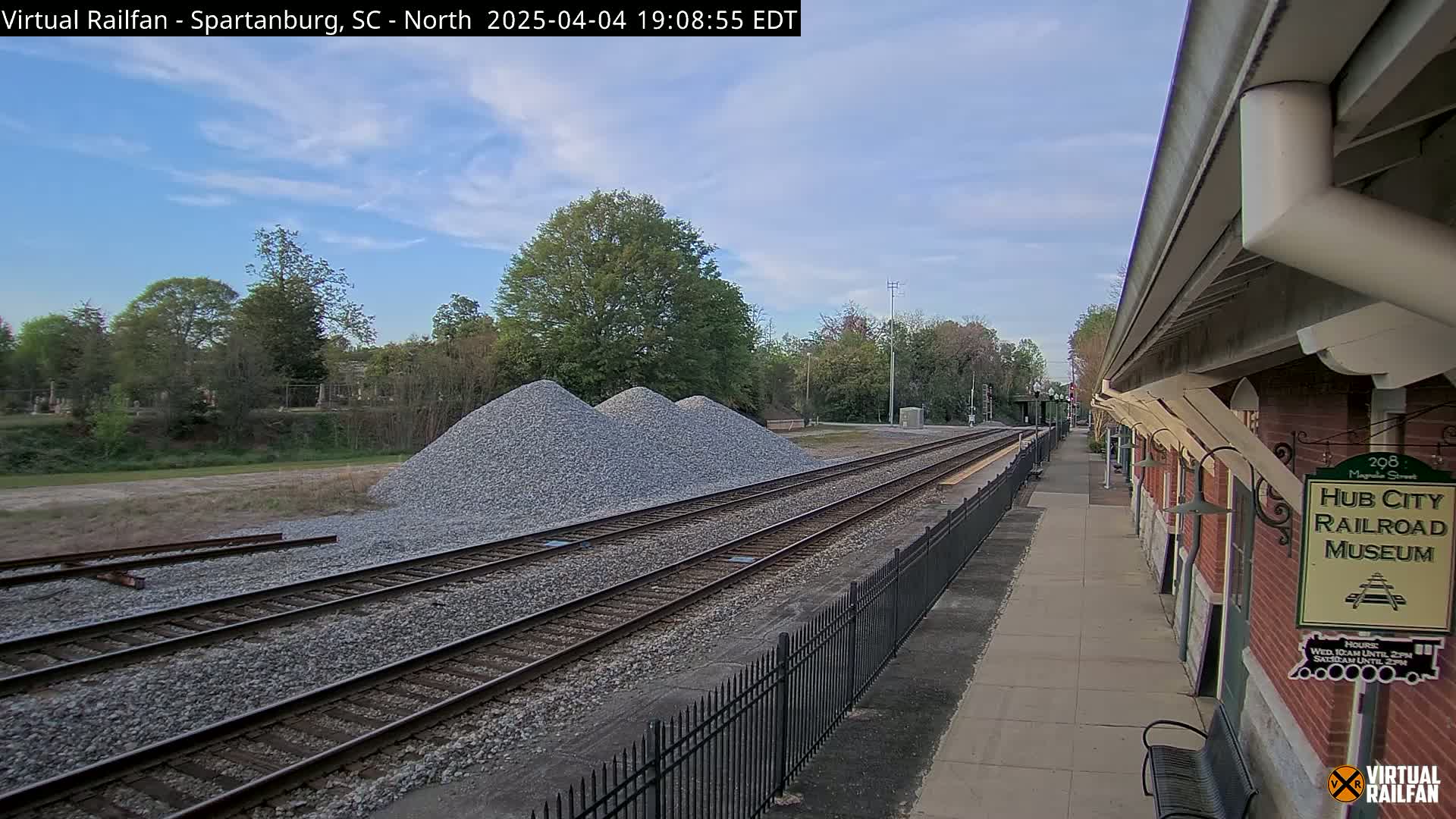 A partly cloudy daytime scene shows railroad tracks next to a brick building, with piles of gravel nearby.