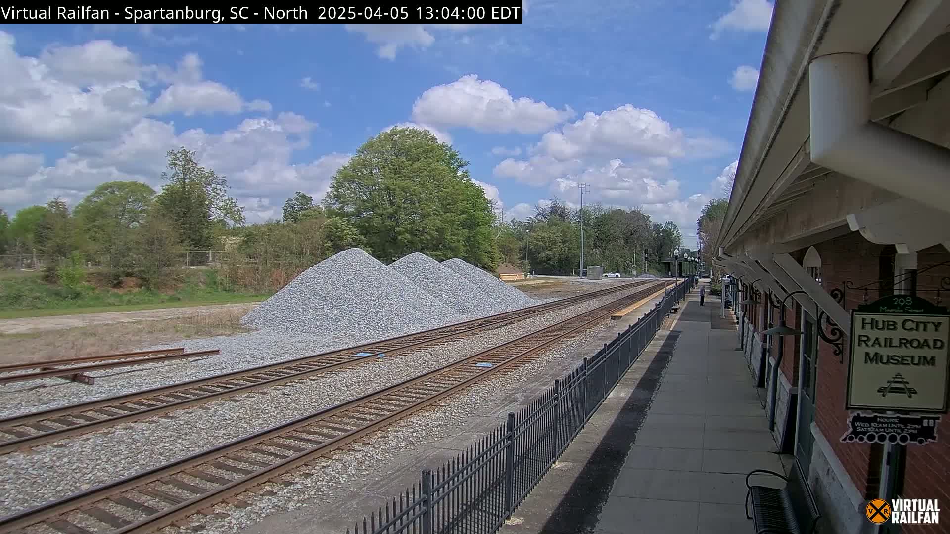 A sunny day shows train tracks next to a brick building and piles of gravel, with trees and vegetation in the background.