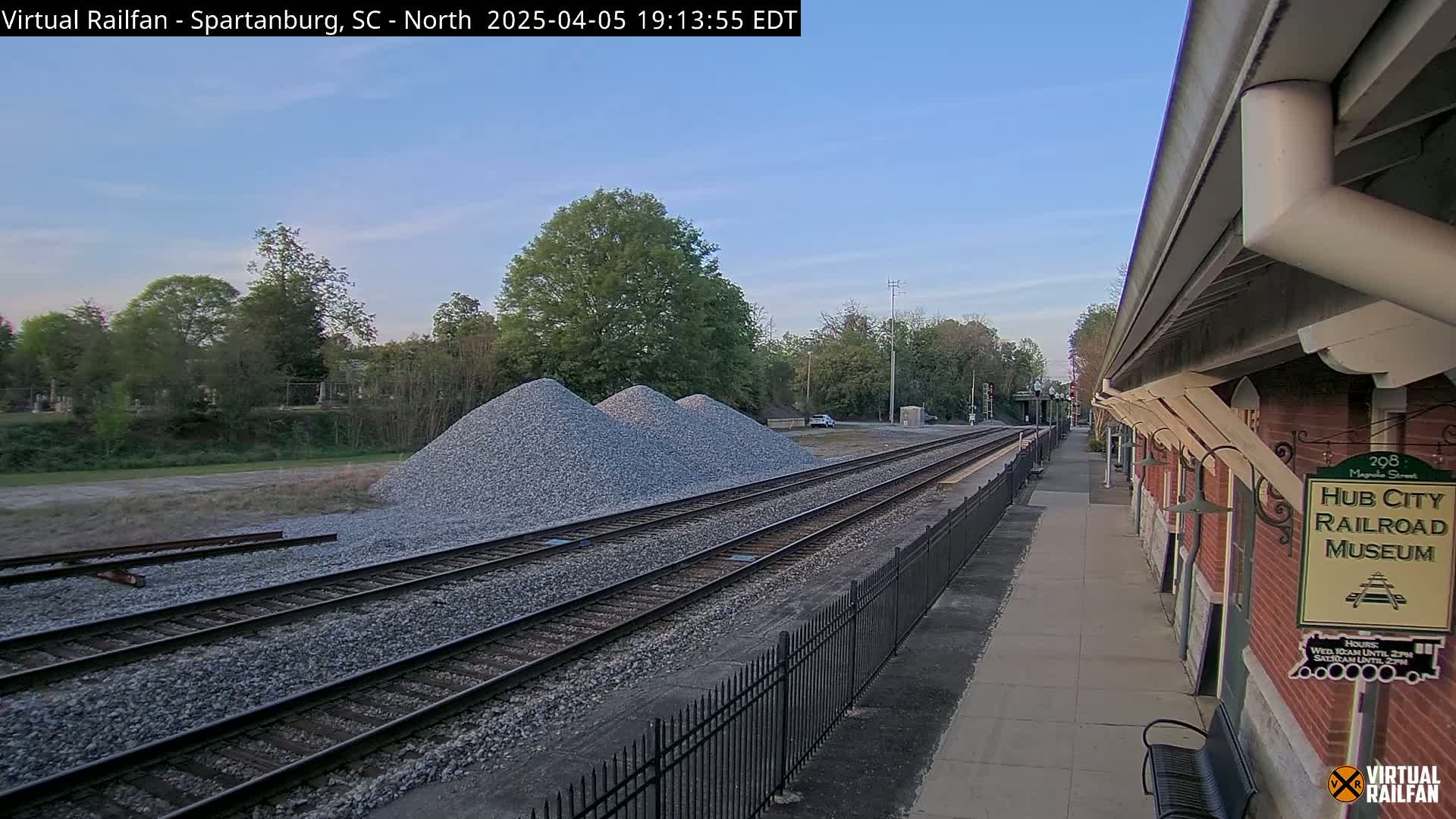 A train station platform with a brick building, railroad tracks, and piles of gravel is visible under a clear, twilight sky.