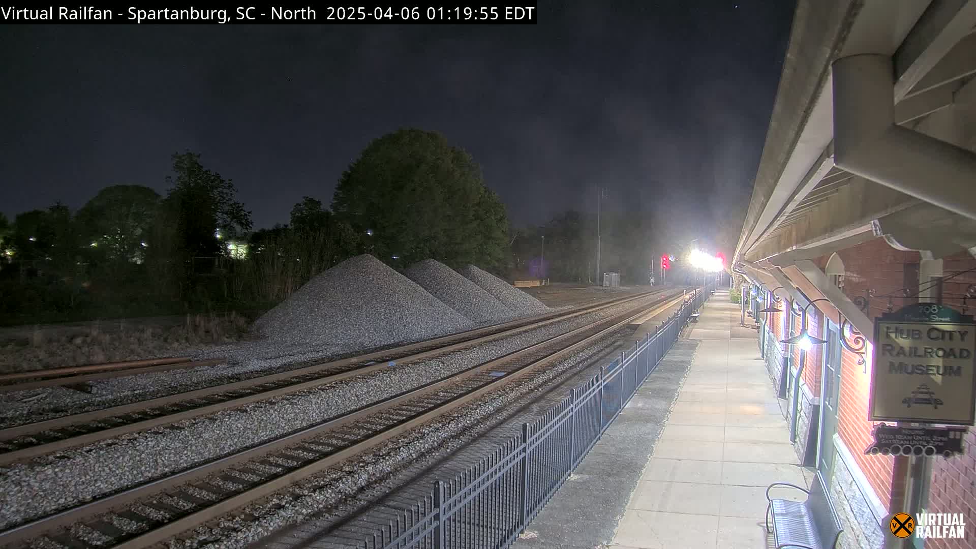 Under a dark night sky, railroad tracks extend from a nighttime train station platform with piles of gravel nearby.