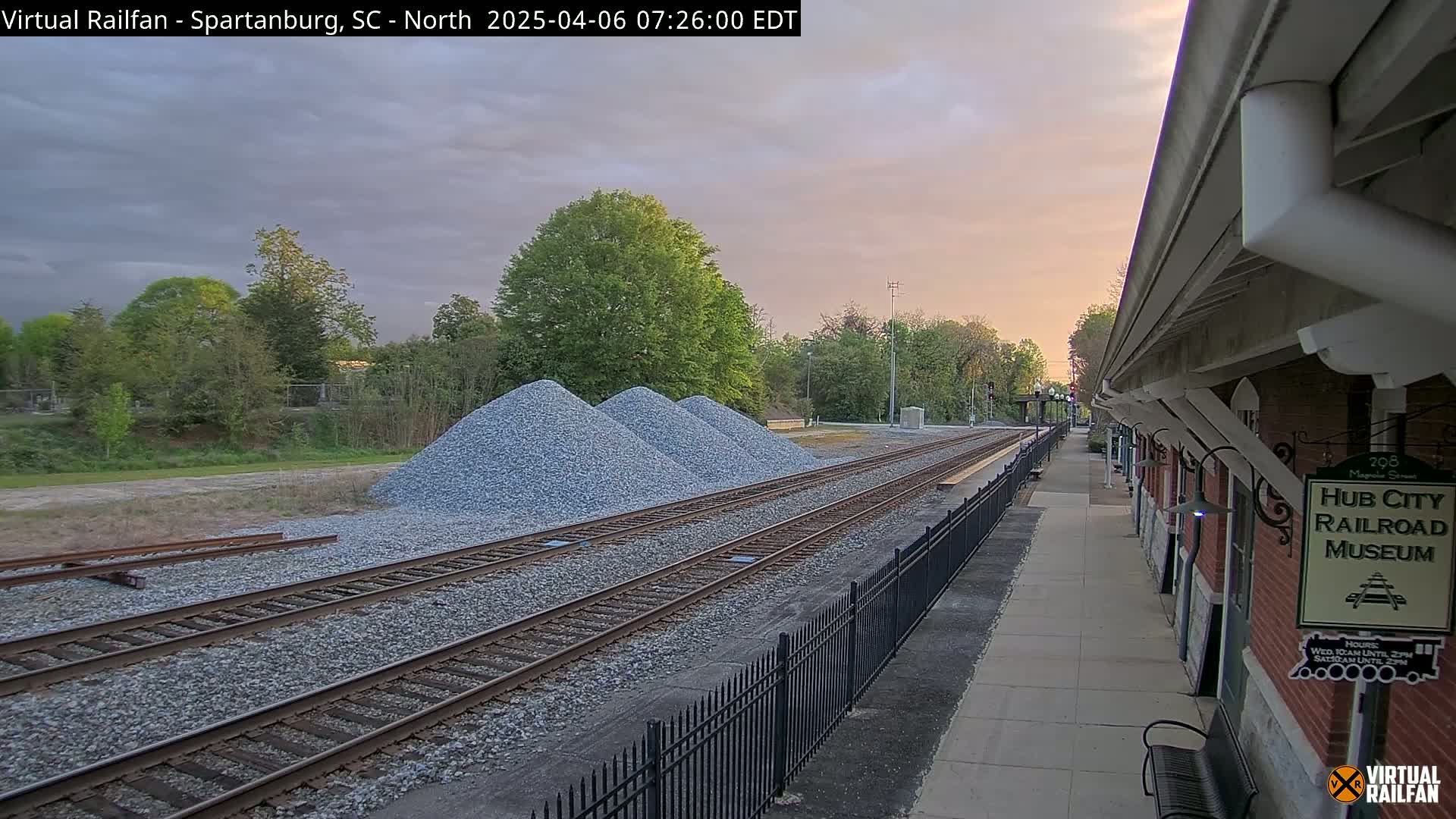 A railroad track with piles of gravel next to it runs alongside a brick railroad museum under a partly cloudy sunset sky.