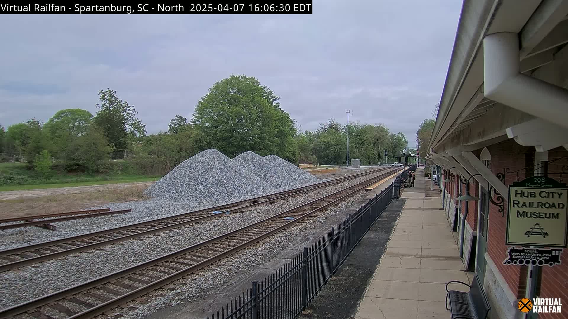 Three piles of gravel sit beside railroad tracks next to a brick building, under a mostly cloudy sky.