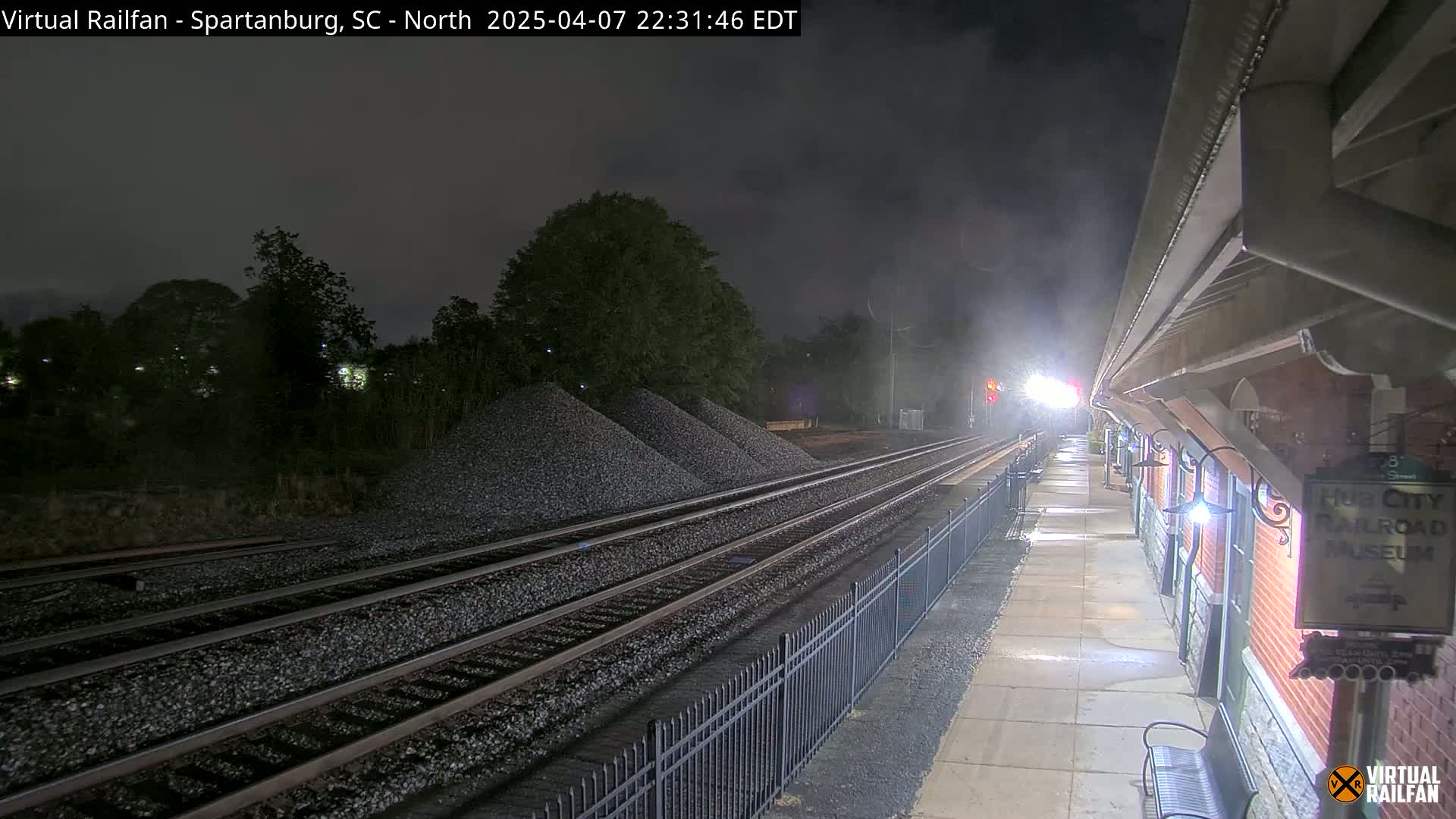 At night, train tracks extend from a railway station platform past piles of gravel under a dark, cloudy sky.