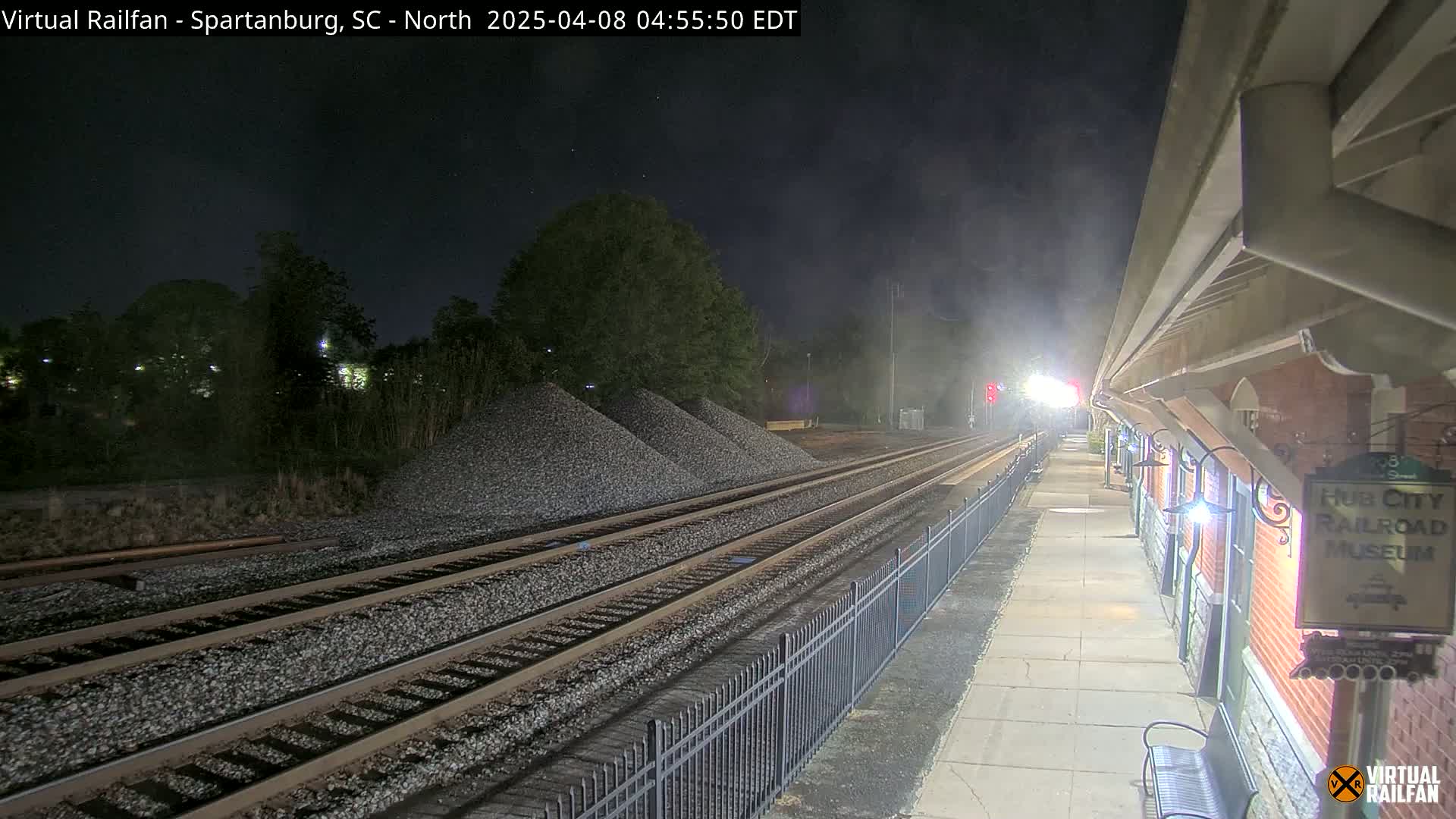 At night, multiple train tracks extend from a railway station platform with a fence separating the tracks and platform, piles of gravel are visible near the tracks.