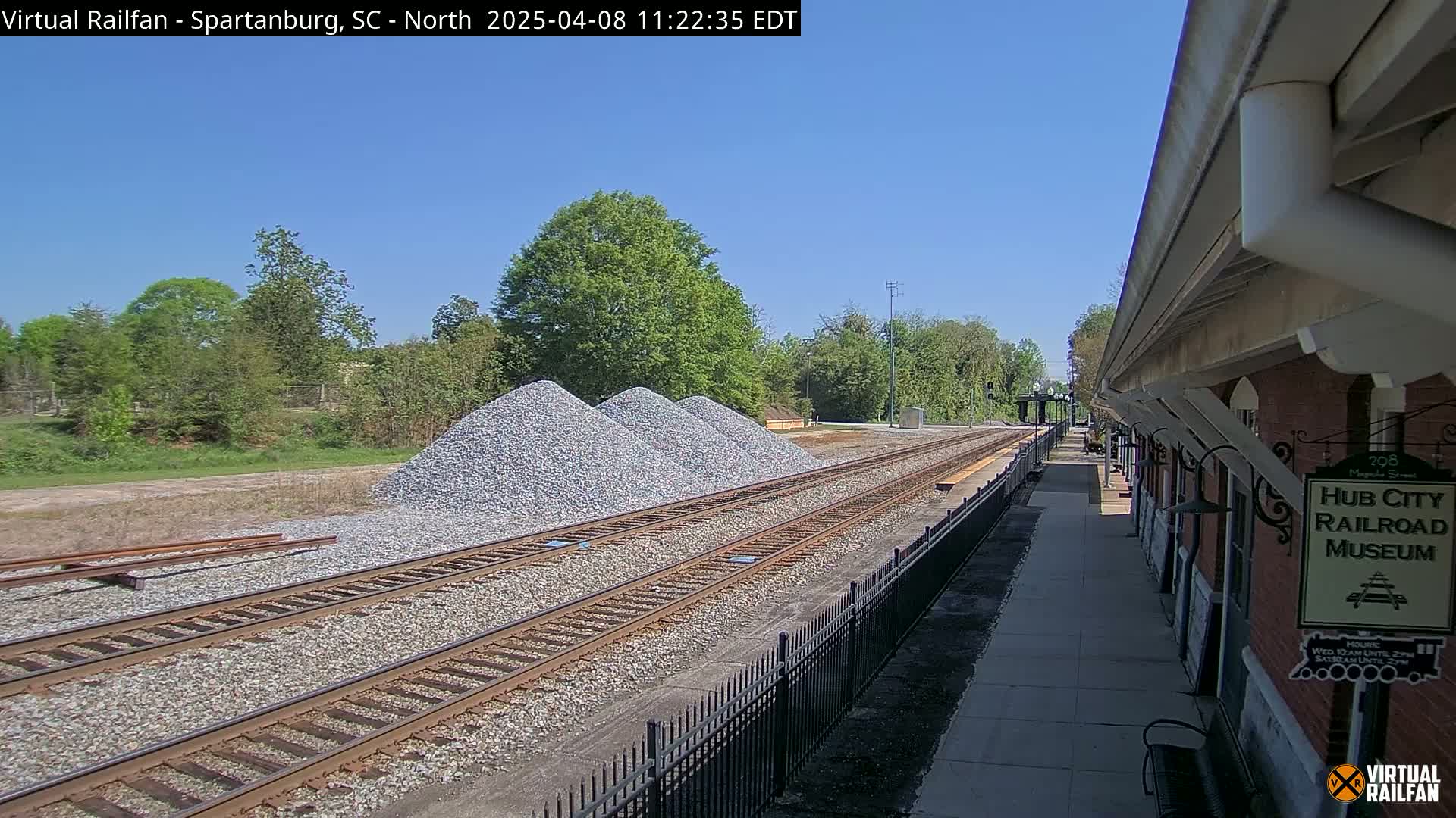 Under a clear blue sky, railroad tracks extend past piles of gravel toward a brick railroad museum.