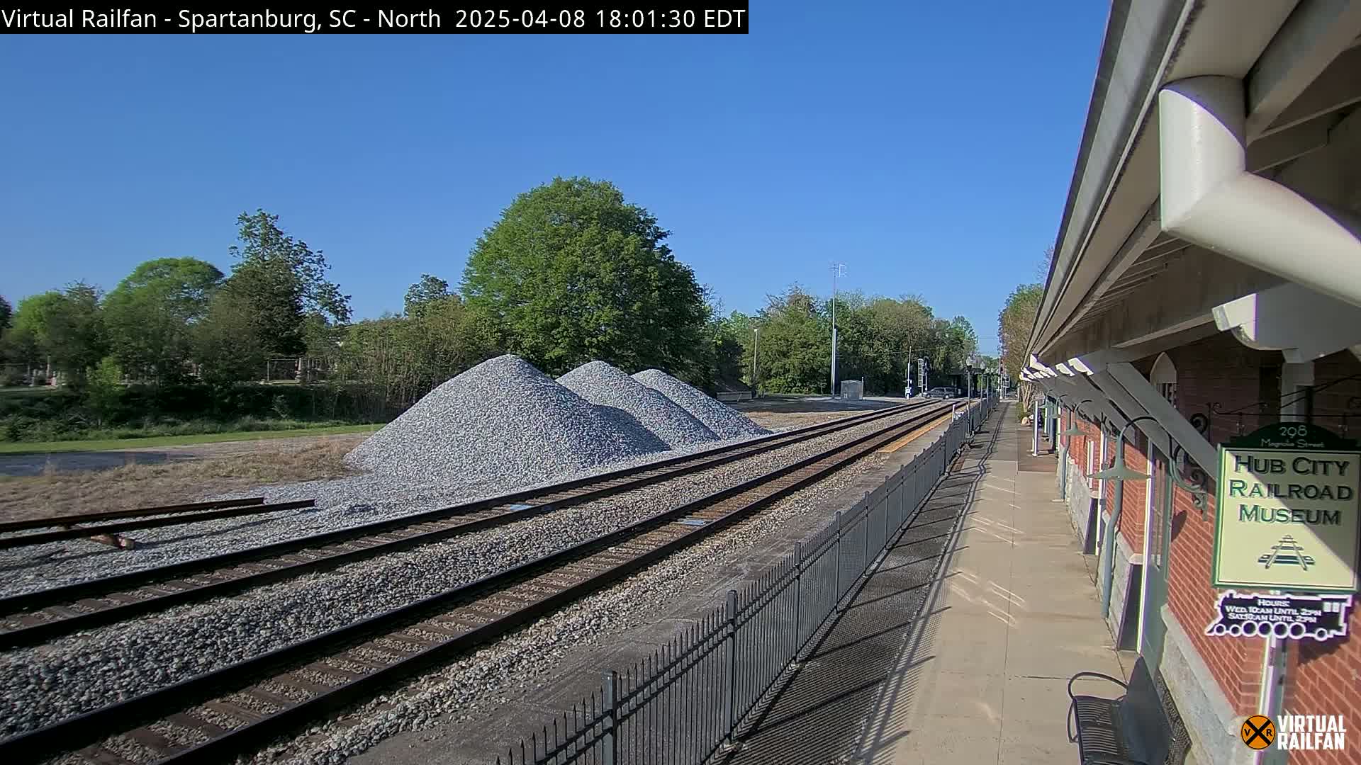 A sunny day reveals railroad tracks with piles of gravel beside them, leading to a brick railroad museum building with a platform and a fence.