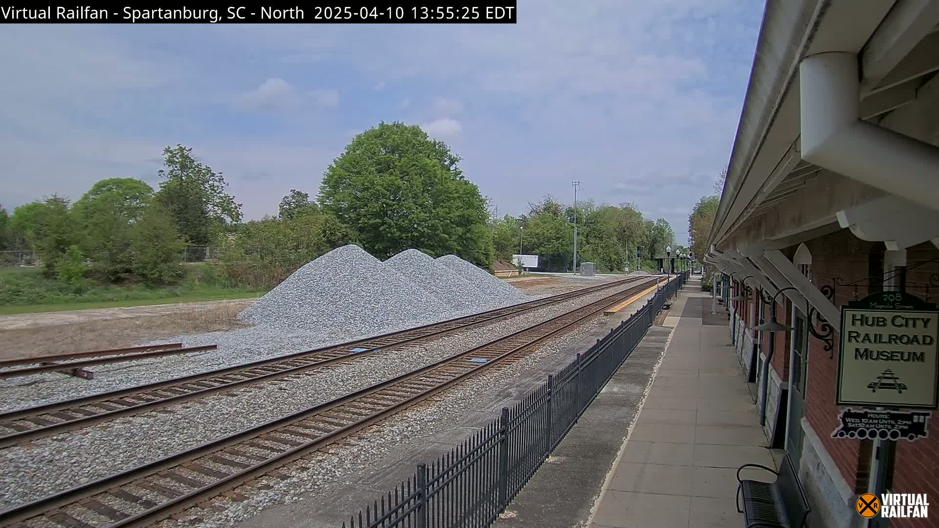 Three piles of gravel sit beside railroad tracks next to a brick building under a partly cloudy sky.