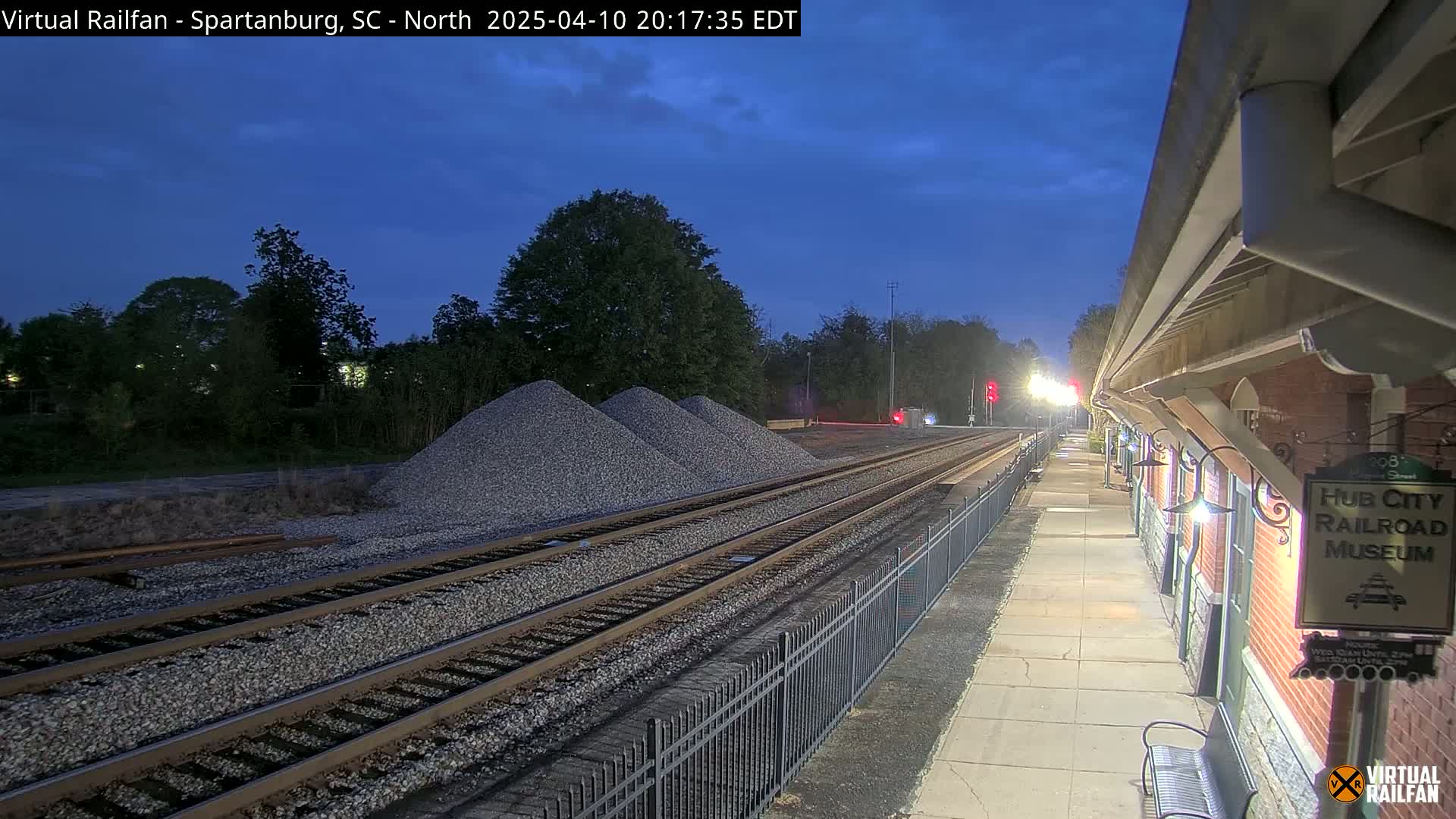 Under a twilight sky, railroad tracks extend from a brick building towards piles of gravel and trees.