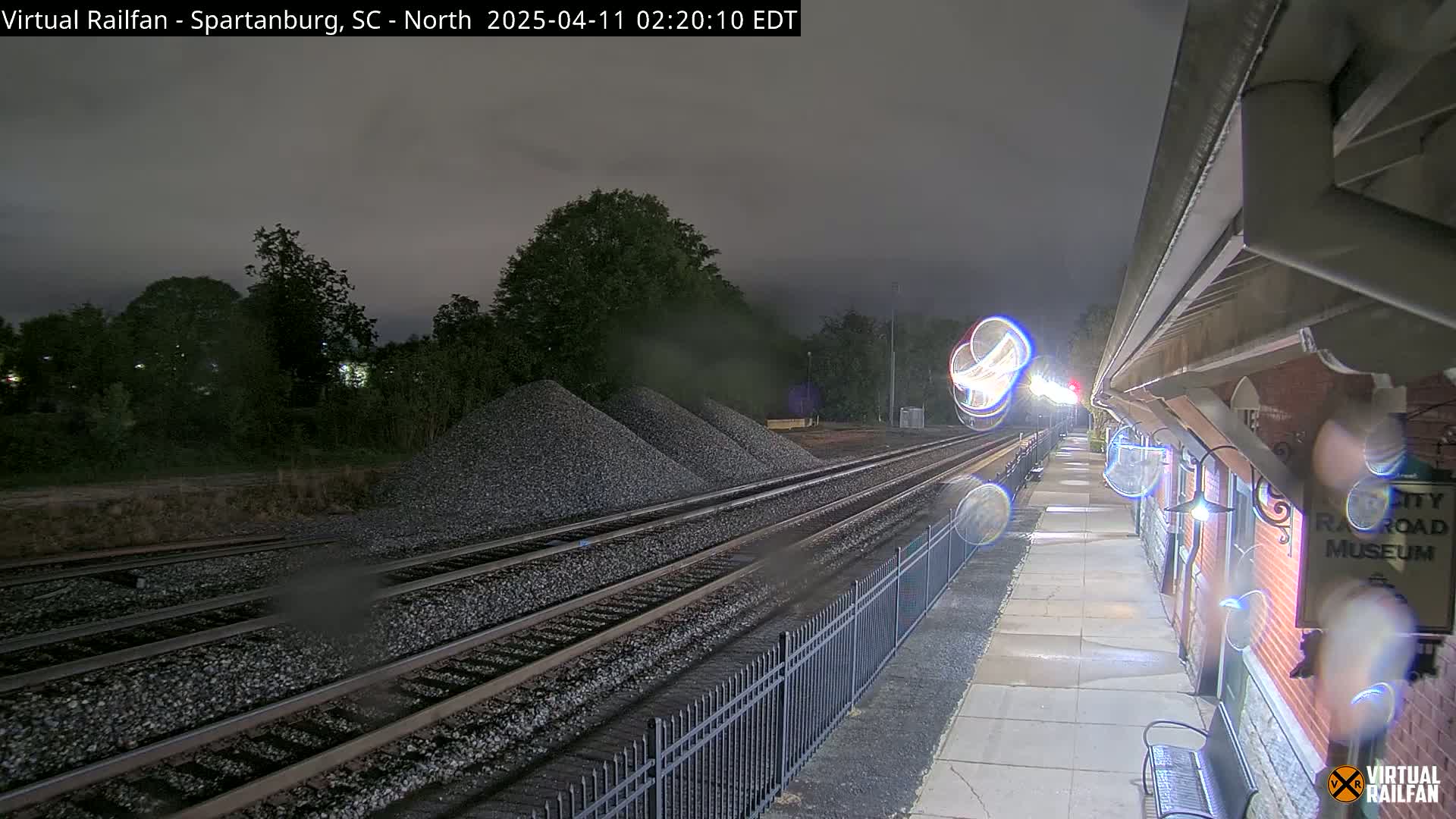 A nighttime view of a train station platform next to railroad tracks and piles of gravel under a dark, possibly rainy sky.