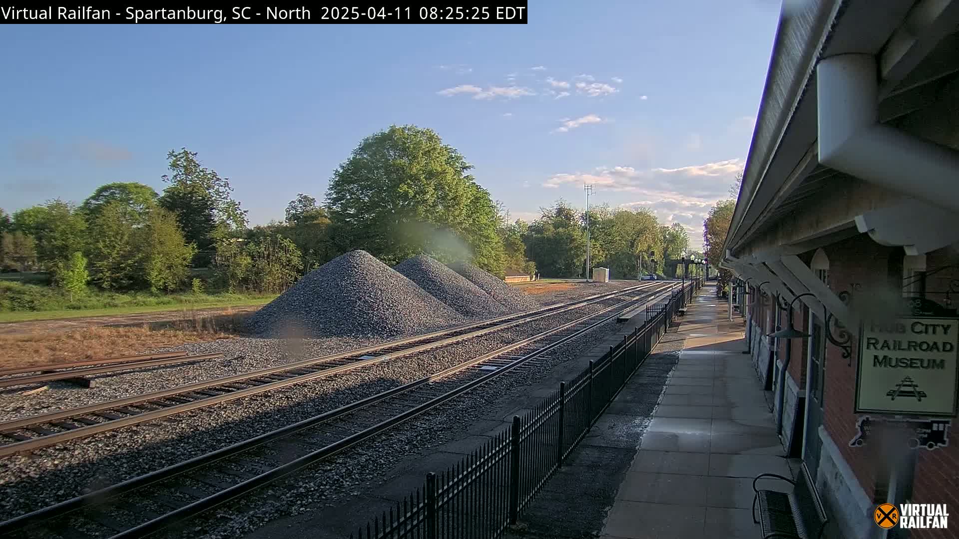 Several piles of gravel sit beside railroad tracks next to a train station platform on a partly cloudy day.