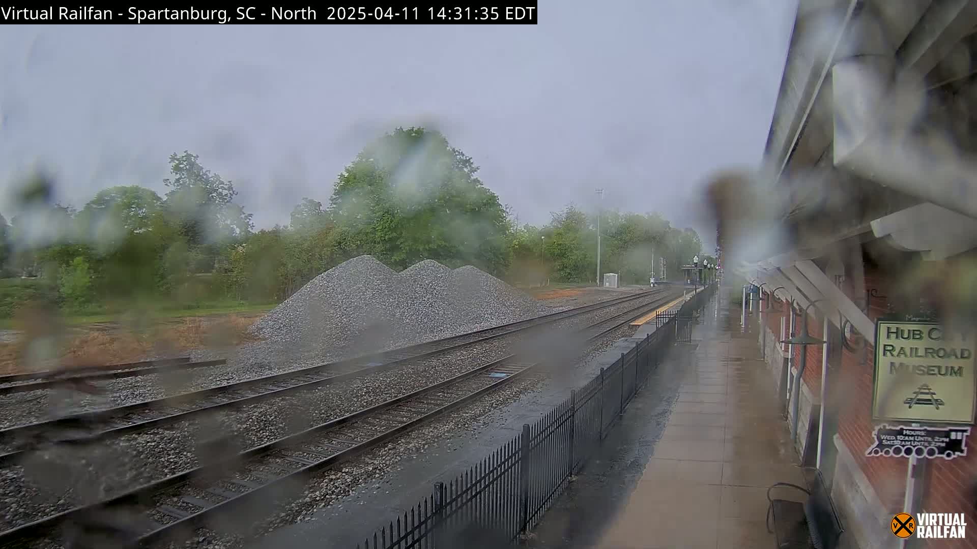 A rainy view of train tracks, a pile of gravel, and a railroad museum platform.
