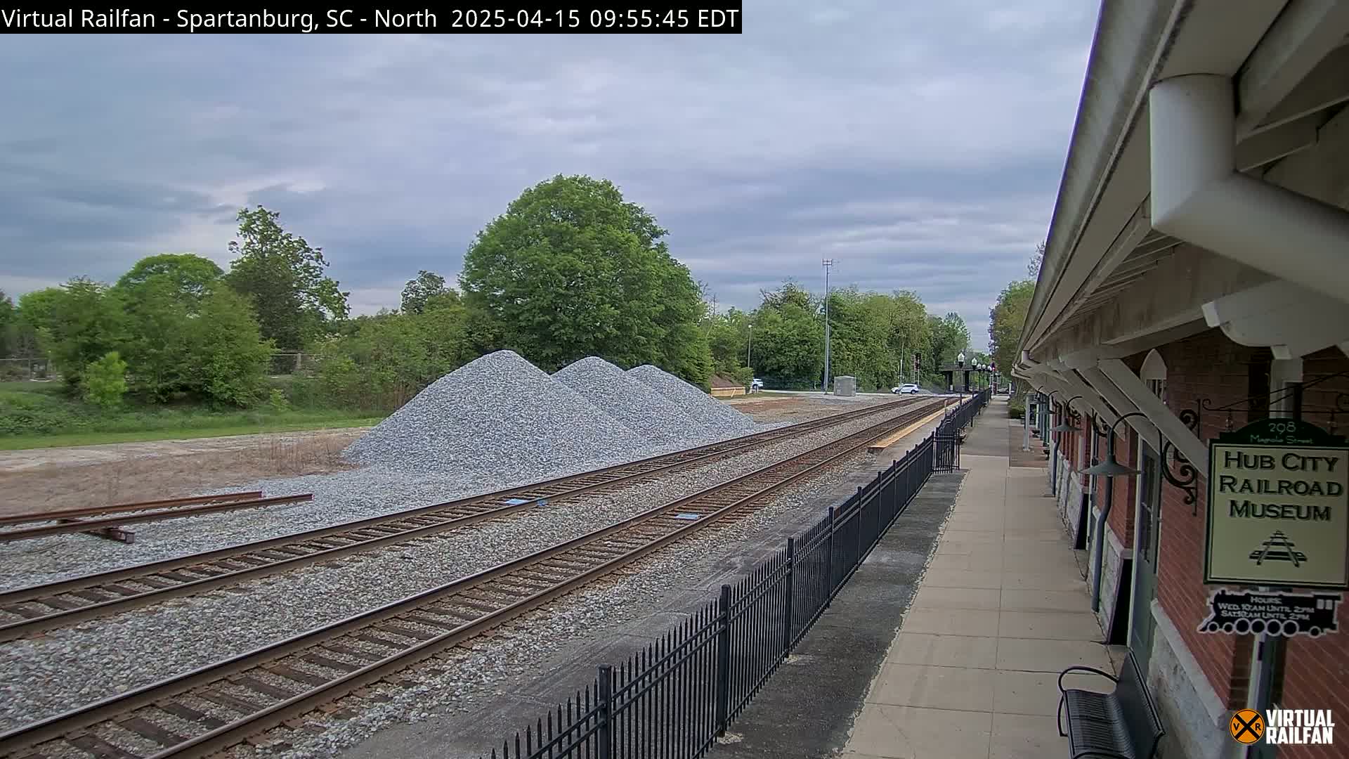 Three piles of gravel sit beside train tracks next to a brick building under a partly cloudy sky.