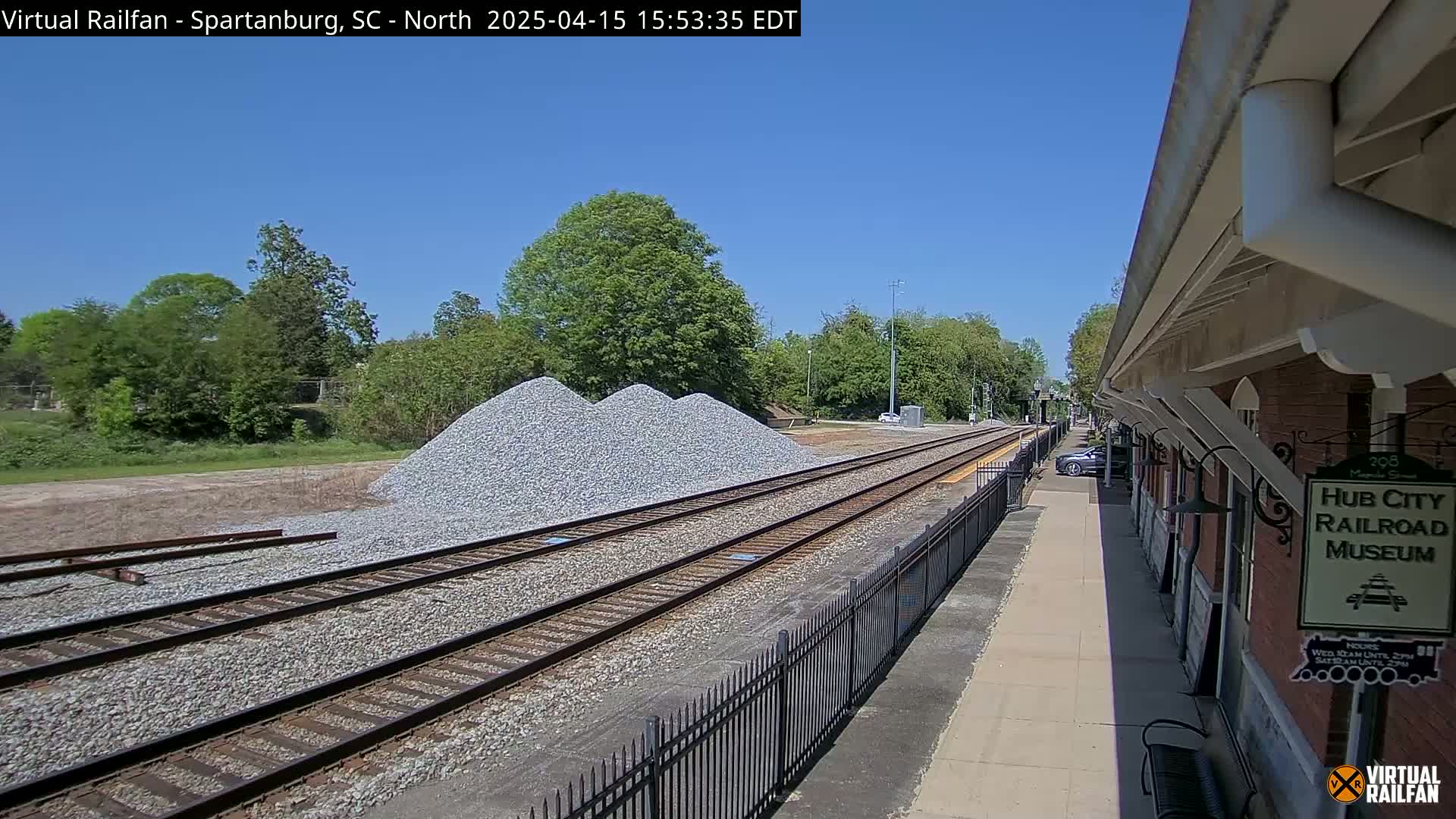 Under a clear blue sky, railroad tracks extend from a train station alongside piles of gravel, with green trees and a paved walkway visible in the background.