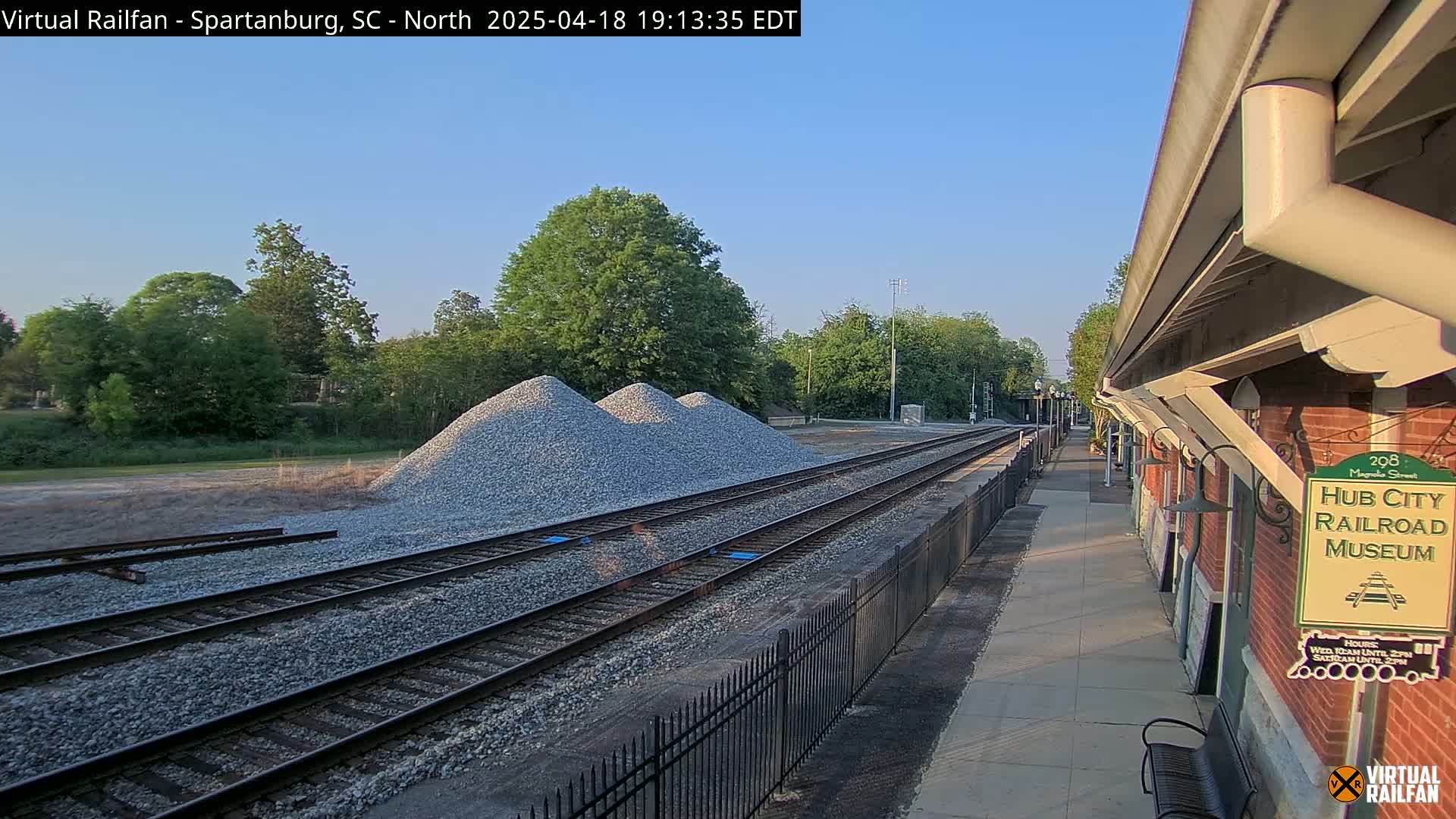 Under a clear sky, railroad tracks extend toward a train station next to piles of gravel, with lush green trees in the background.